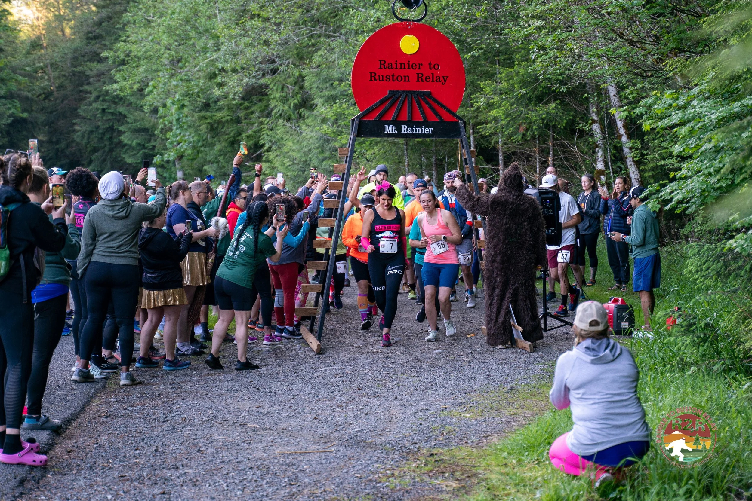 Participants crossing the starting line of a trail race called 'Rainier to Ruston Relay' in a forested area, with a mascot in a bear costume and spectators taking photos.