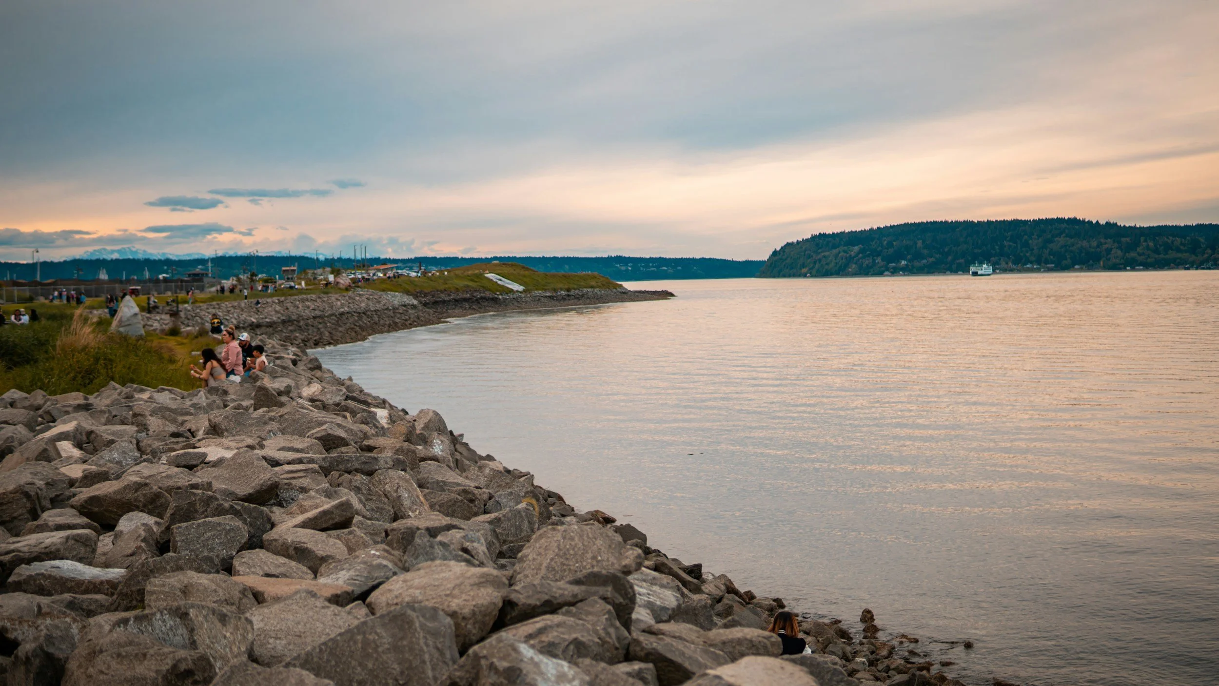 People sitting and walking along a rocky shoreline at sunset, with a view of water and distant hills under a cloudy sky.