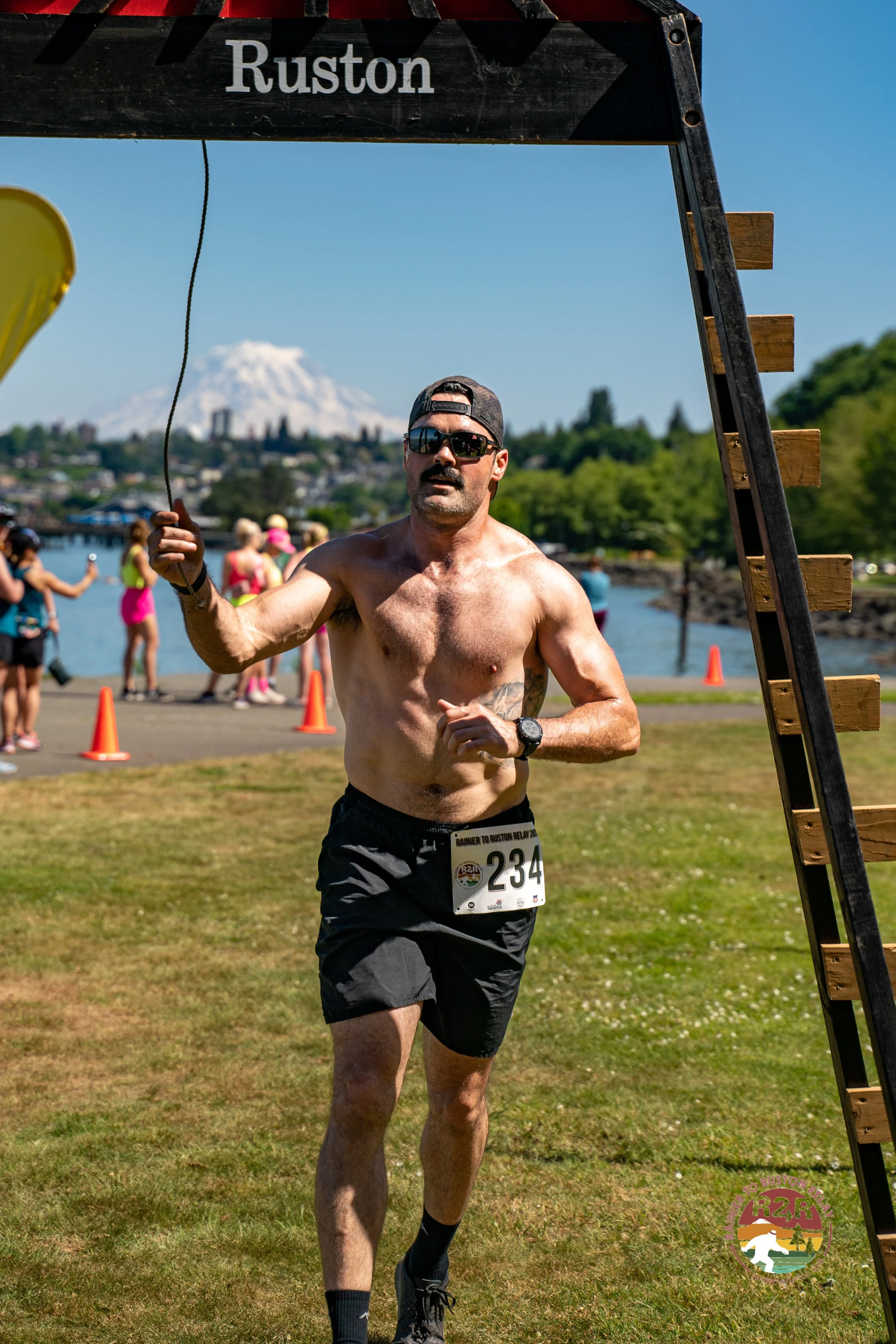 A shirtless man running a race wearing black shorts, sunglasses, and a cap, with a race bib numbered 234, crossing the finish line outdoors on a sunny day, with other runners and a scenic background including a body of water, trees, and a snow-capped