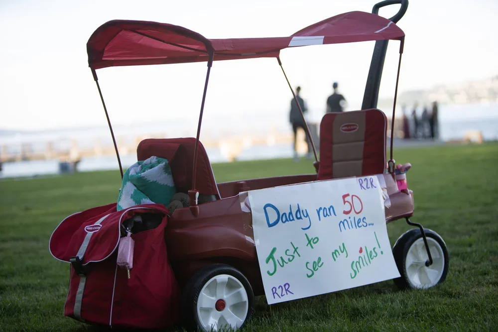 A red pedal-powered toy car with a canopy, parked on grass near water. It has a sign that reads, 'Daddy ran 50 miles... Just to see my smiles!'