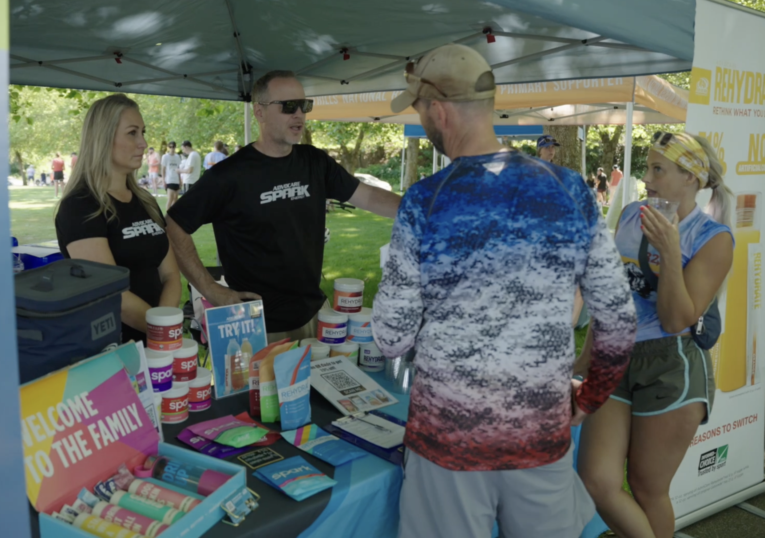 People at an outdoor booth promoting ReHydrate products, with displays of containers and pamphlets, under a tent on a park day.