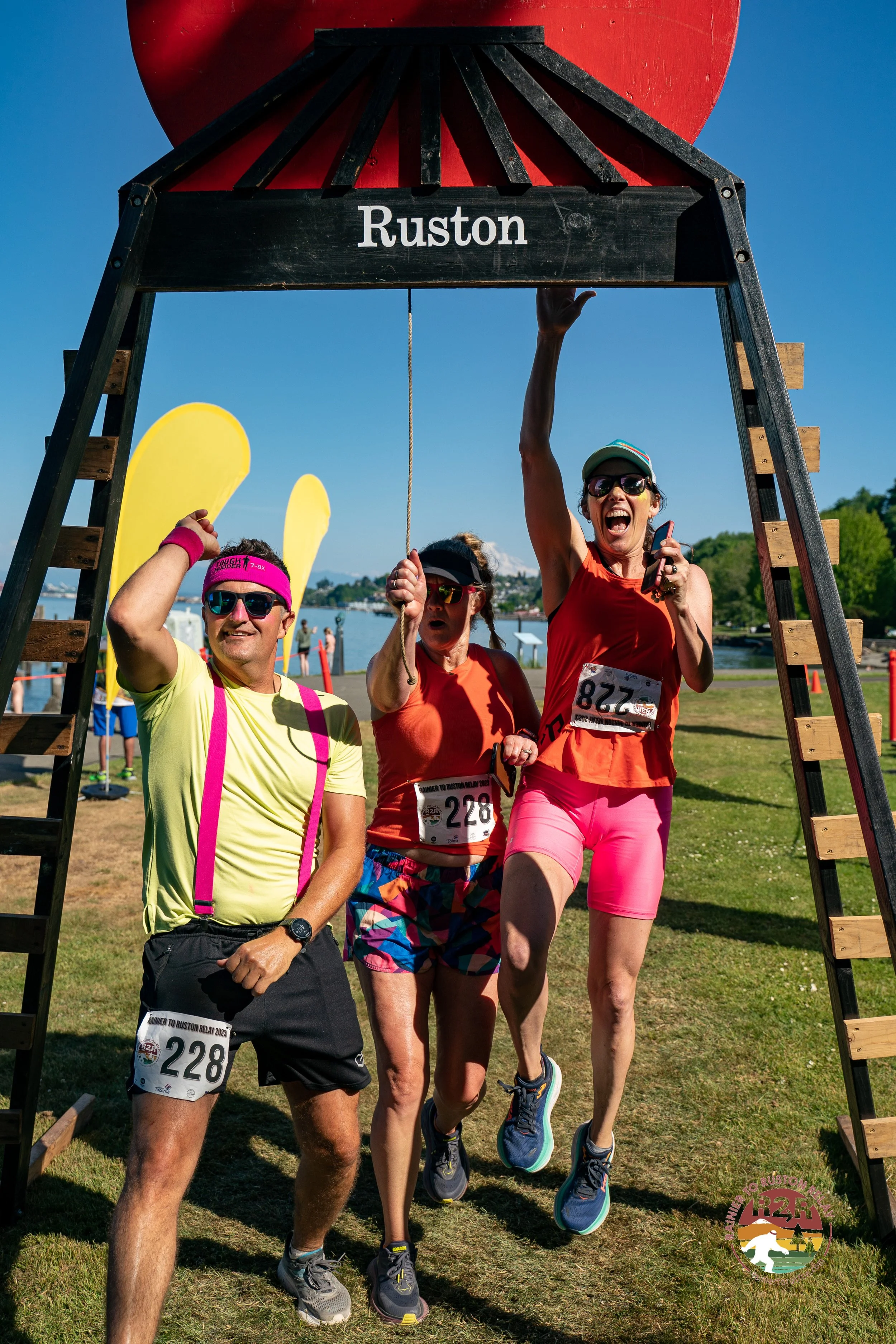 Three people celebrating at the finish line of a race in Ruston, holding up a black wooden sign with 'Ruston' written on it. The man on the left is wearing a yellow shirt, black shorts, pink suspenders, sunglasses, and a pink headband. The woman in the center is wearing an orange tank top, colorful shorts, sunglasses, and a race bib. The woman on the right is wearing a red tank top, pink shorts, sunglasses, a cap, and is holding a phone, jumping in excitement.