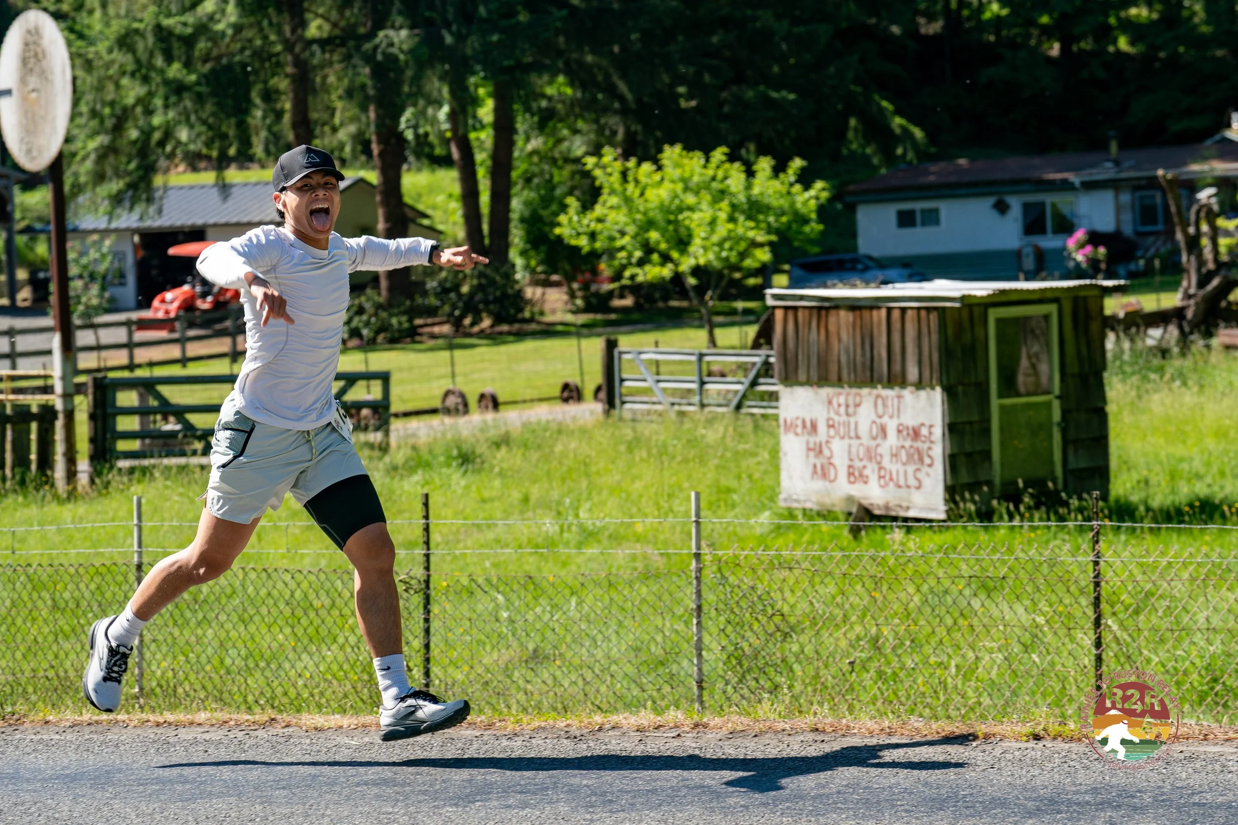 A young man running outdoors on a sunny day, wearing athletic clothes and a cap, with a surprised or excited expression, in a rural area with green grass, trees, a wooden fence, and a small building in the background.
