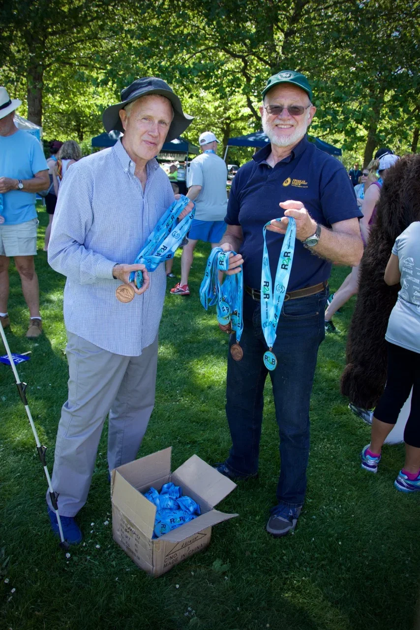 Two men standing outdoors at a park, holding medals with blue ribbons, after a race event. One man is wearing a wide-brimmed hat and light-colored clothes, while the other is wearing a cap, sunglasses, and a dark shirt. There is a box on the ground filled with more medals, with several people and trees in the background.
