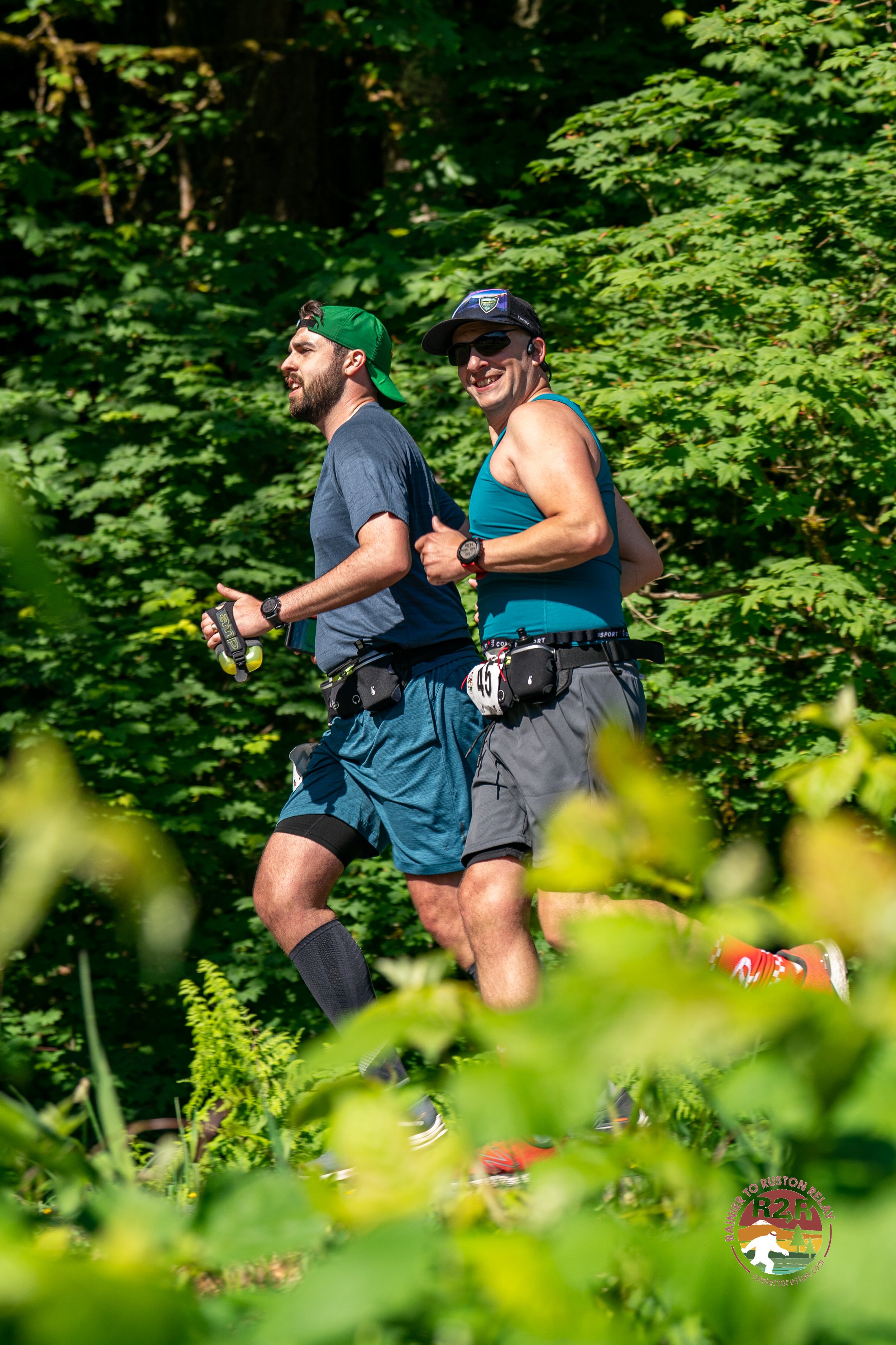 Two men running outdoors on a trail surrounded by lush green trees, wearing athletic gear and water bottles, smiling and enjoying a sunny day.