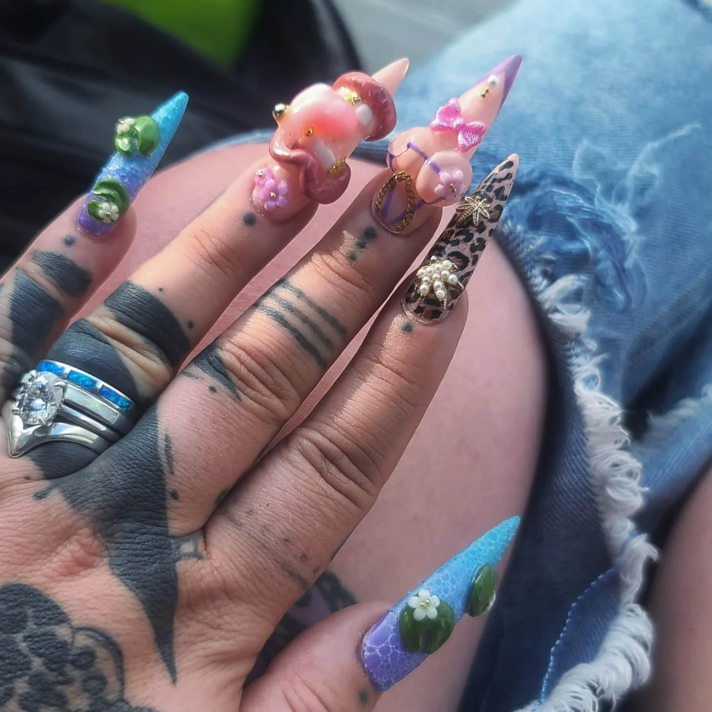 Close-up of a hand with decorated long, pointed nails featuring various 3D embellishments and designs, including flowers, a leopard print, and a star-shaped charm, with rings on the fingers and tattoos visible.