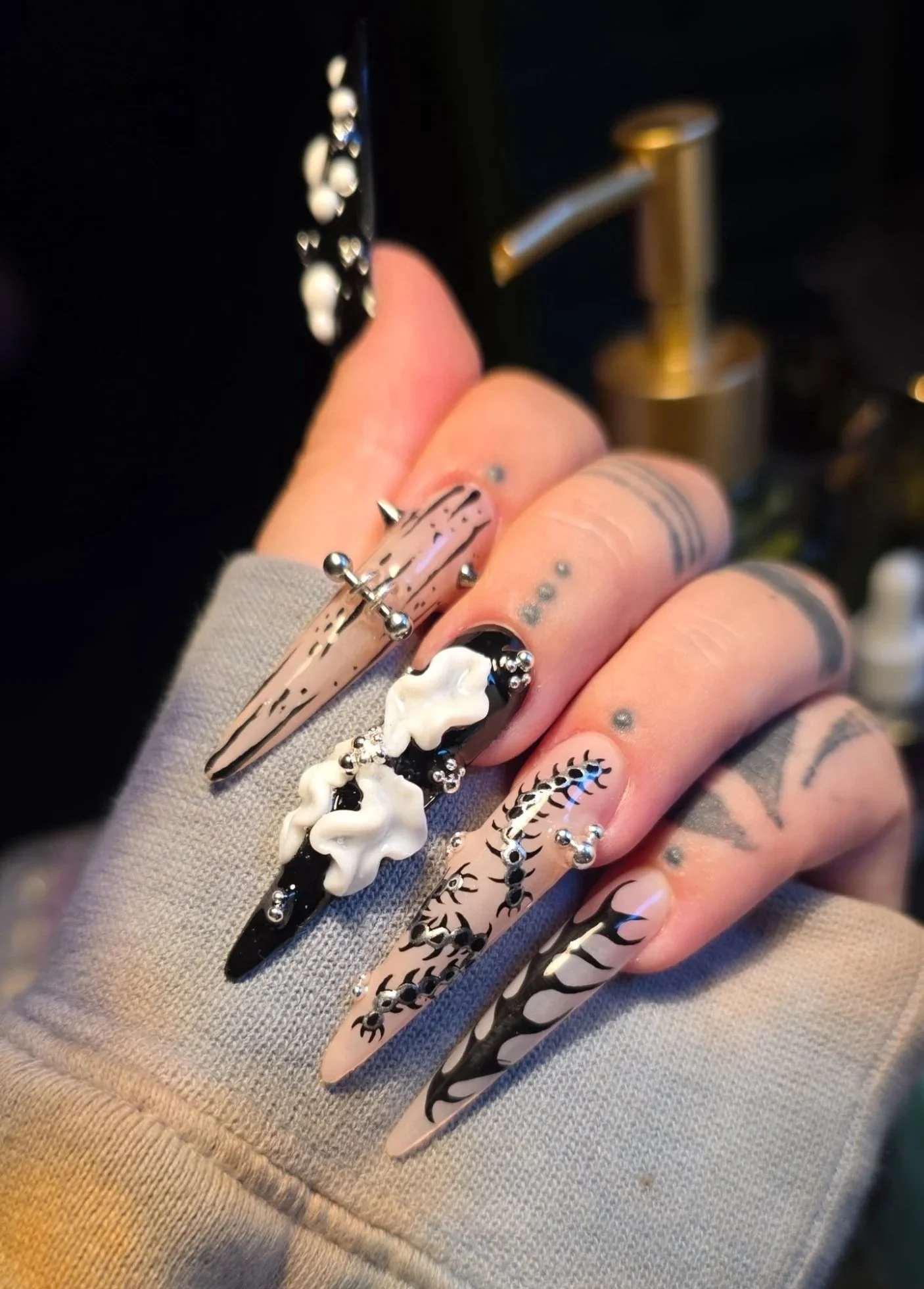 A hand with long, coffin-shaped nails painted in a black and nude design, adorned with 3D white cloud-like embellishments and small silver studs, with various black tattoo designs on the fingers.