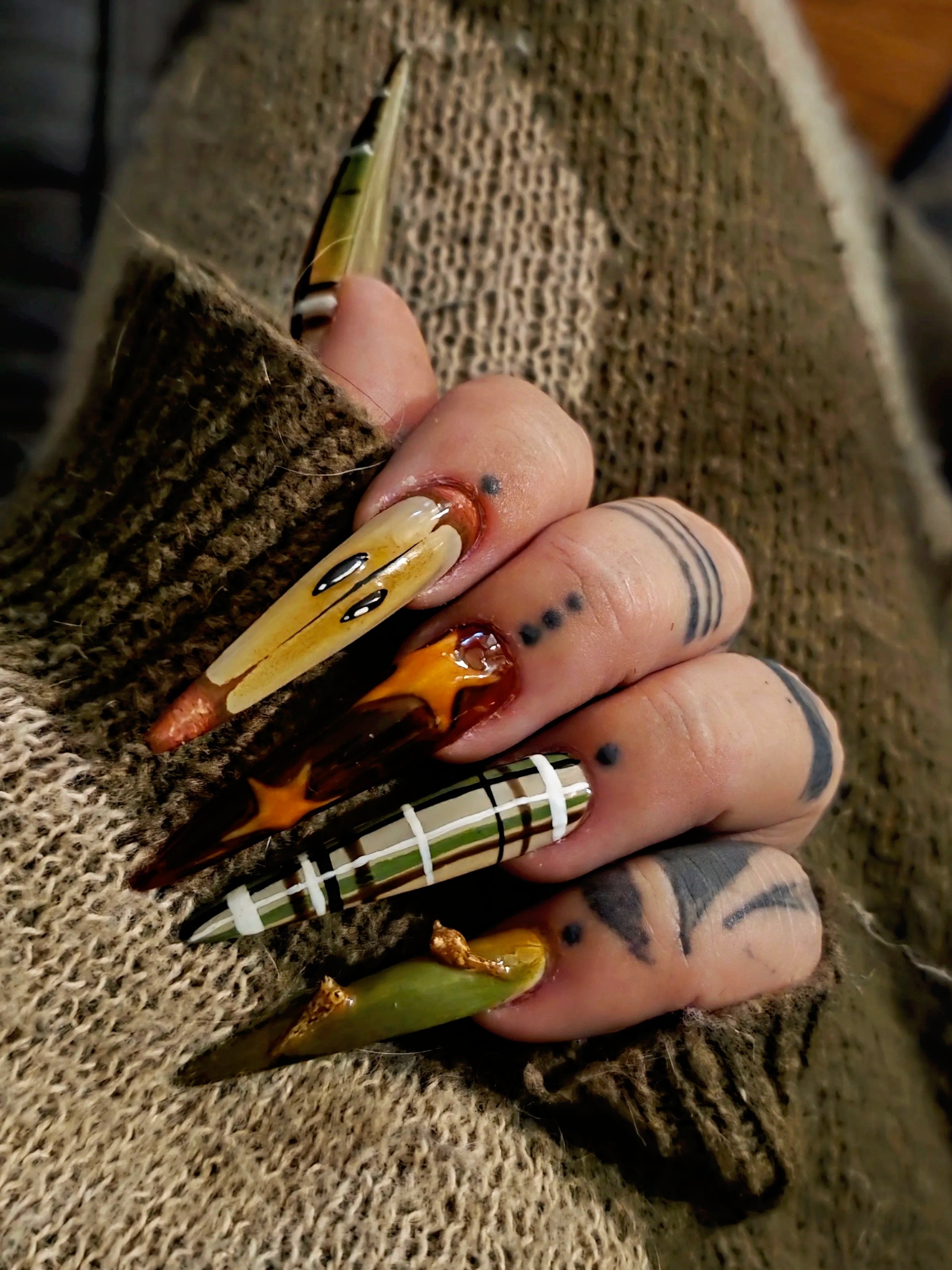 Close-up of a hand with long, decorated acrylic nails, tattoos, and a brown knitted sweater, resting on a textured fabric surface.