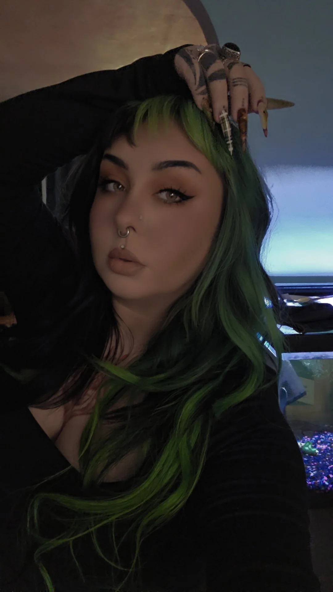 Woman with green and black hair taking a selfie indoors, with aquarium in the background.