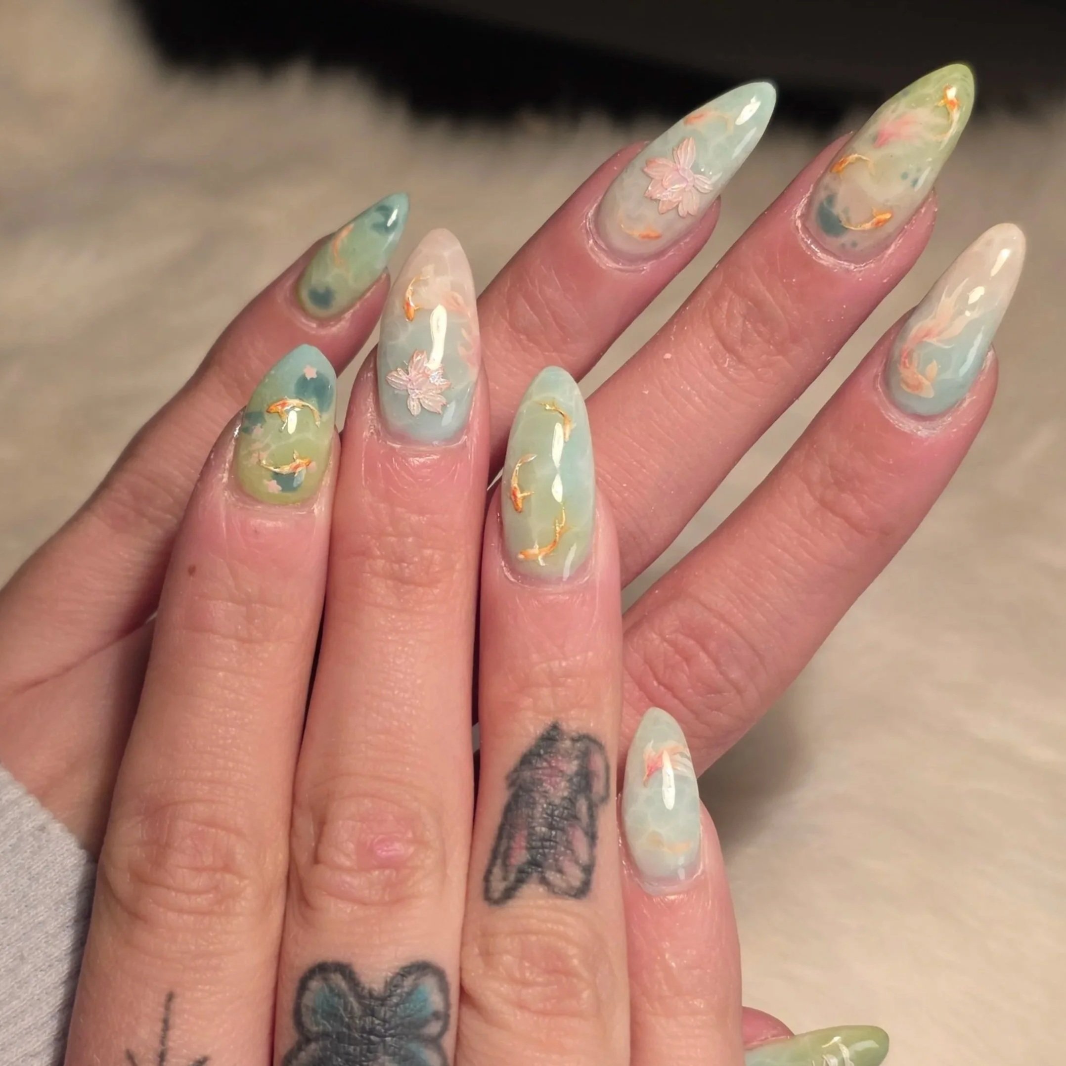 Close-up of a hand with long, colorful, marble-patterned nails with floral and gold accents. The hand has tattoos on the fingers, including a butterfly and a ghost.