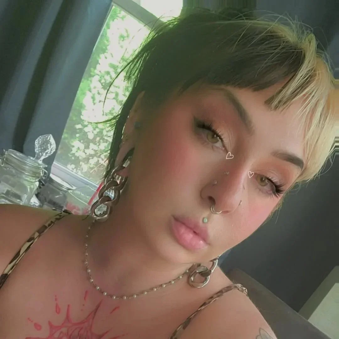 A young woman with short blond and black hair, multiple facial piercings, and makeup featuring natural tones and winged eyeliner. She is wearing large silver hoop earrings, a string of pearls, and a necklace. The background shows a window with green trees outside.