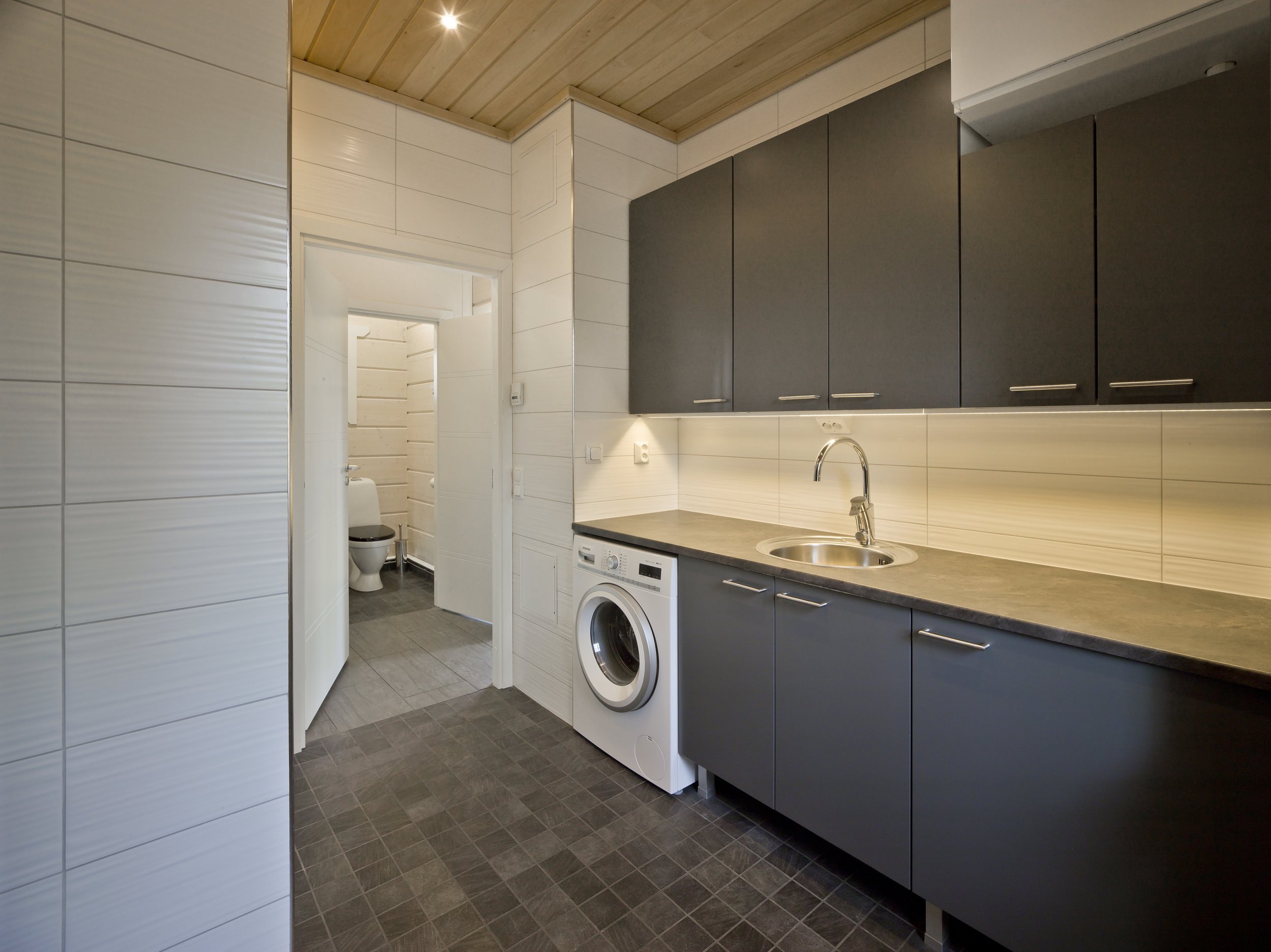 A modern kitchen with dark gray cabinets, a stone countertop, a stainless steel sink, and a washing machine. There is a doorway leading to a small bathroom with a toilet visible. The walls are tiled in white, and the ceiling is wood-paneled.