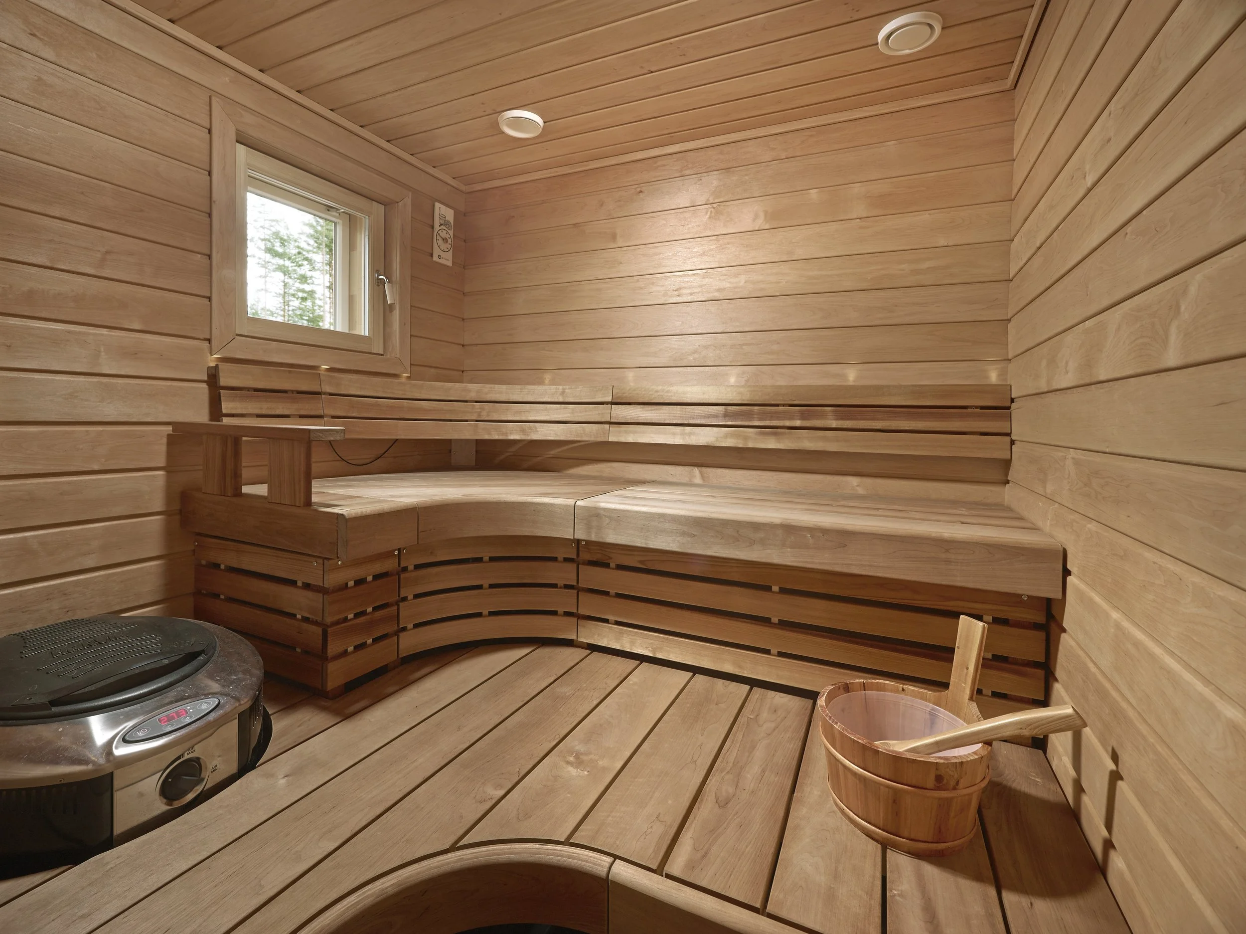 Wooden sauna interior with benches, small window, sauna bucket with ladle, and a heater.