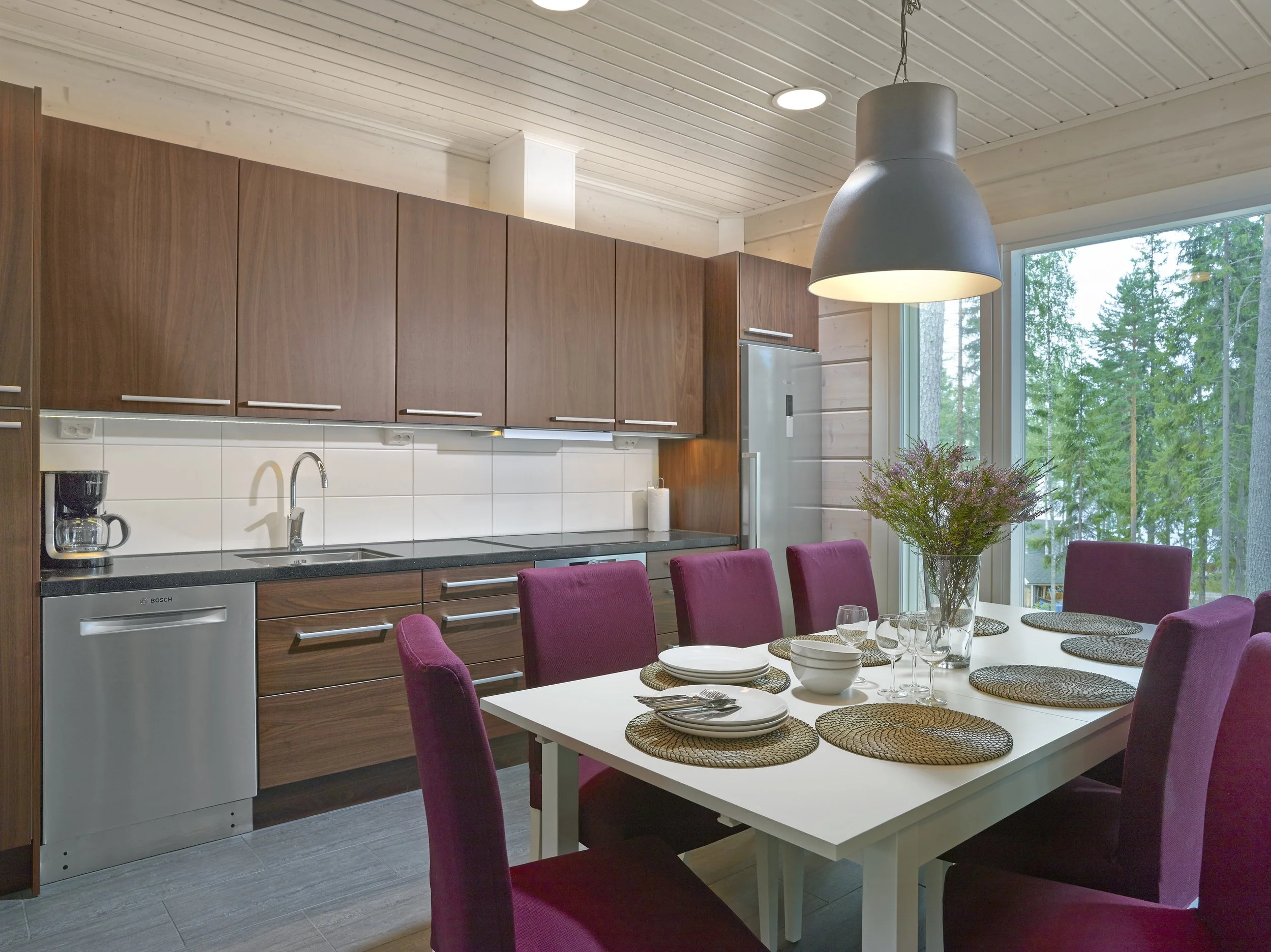 A modern kitchen and dining area with wooden cabinets, a white table set with plates and glasses, purple chairs, a large window showing trees outside, and a hanging light fixture.