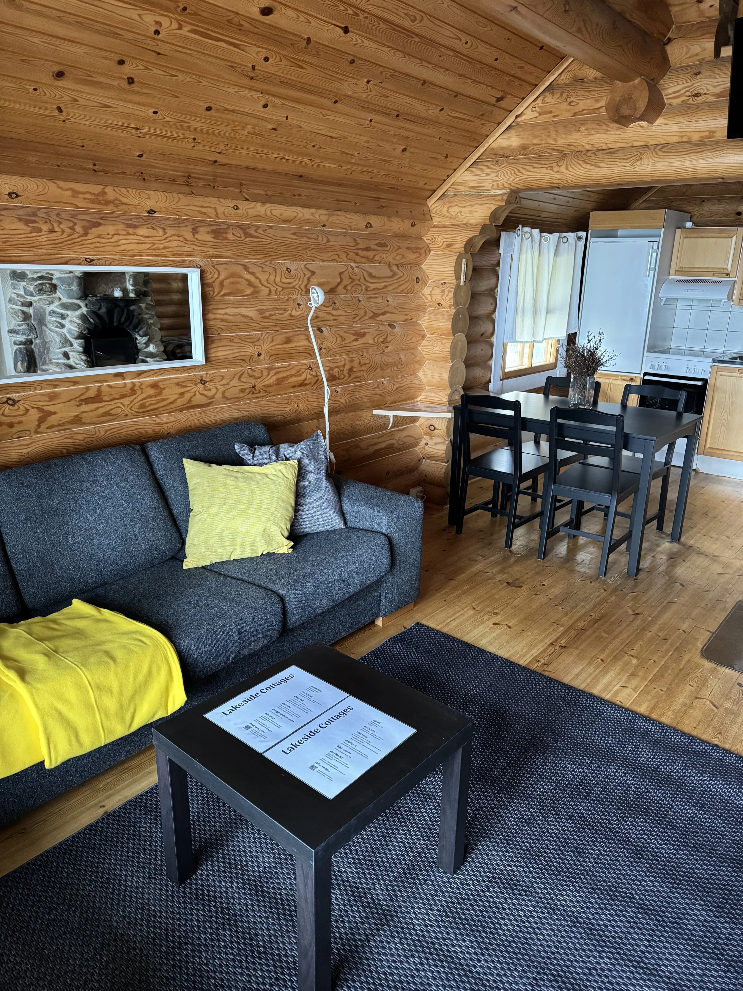 Kuva kodikkaan mökin olohuoneesta, jossa on puiset seinät ja katto, tummanharmaa sohva keltaisilla ja harmailla tyynyillä, pieni musta sohvapöytä, jolla on lehti, ruokapöytä kuudella mustalla tuolilla, ikkuna valkoisilla verhoilla sekä keittiöalue, jossa on