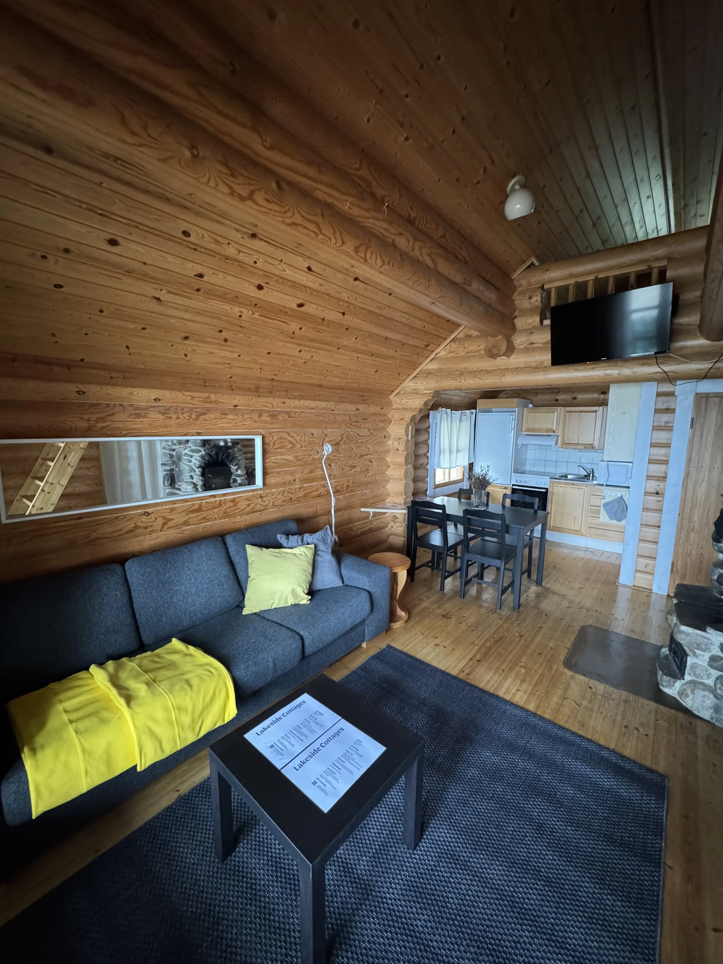 Hirsimökin olohuoneen sisustus: harmaa sohva, musta sohvapöytä, keltainen viltti, puiset seinät ja katto, televisio, keittiönurkkaus sekä ruokapöytä ja tuolit.