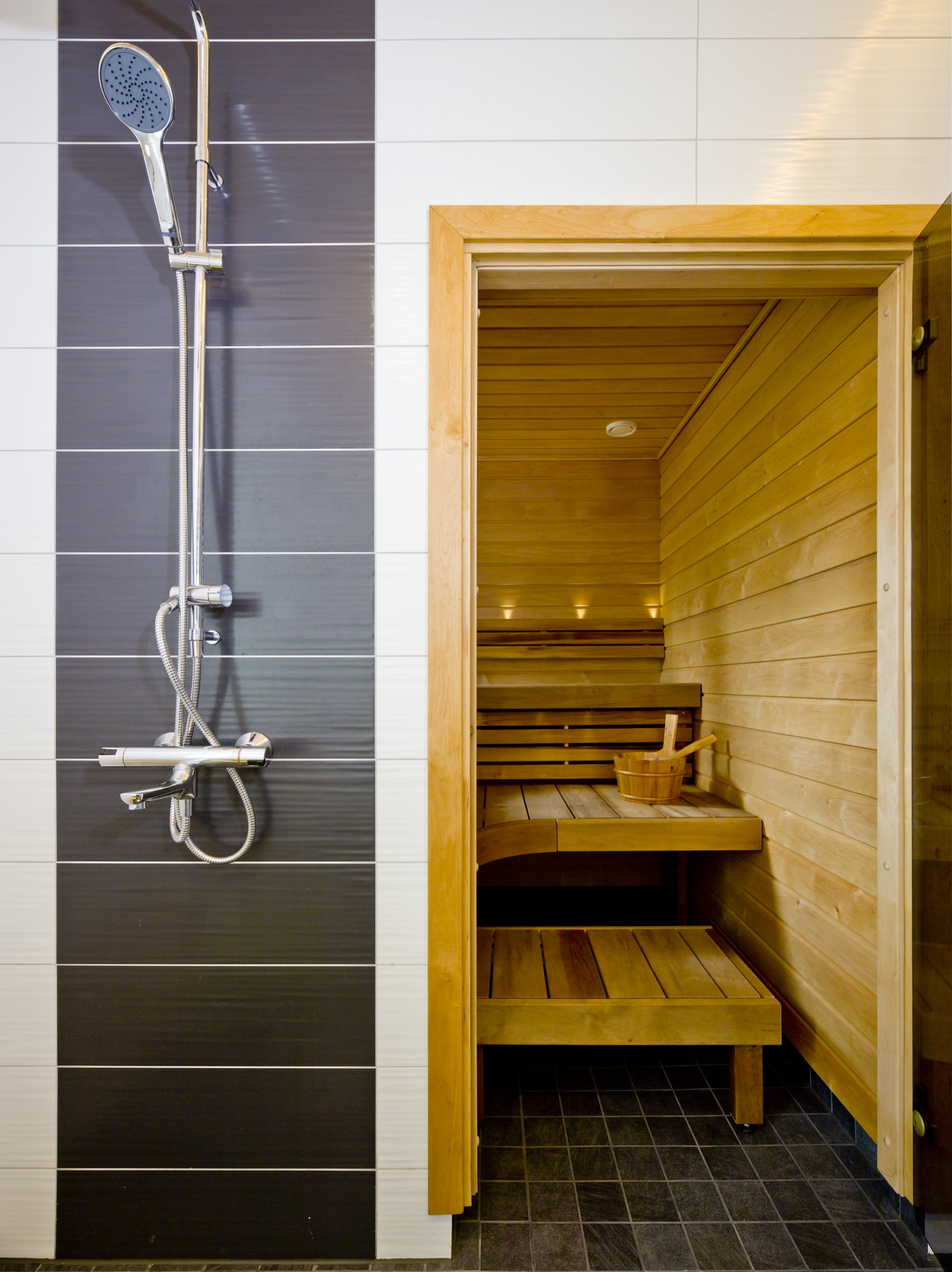 Shower area with a black and white tiled wall and a wooden sauna or steam room with benches and a bucket inside.