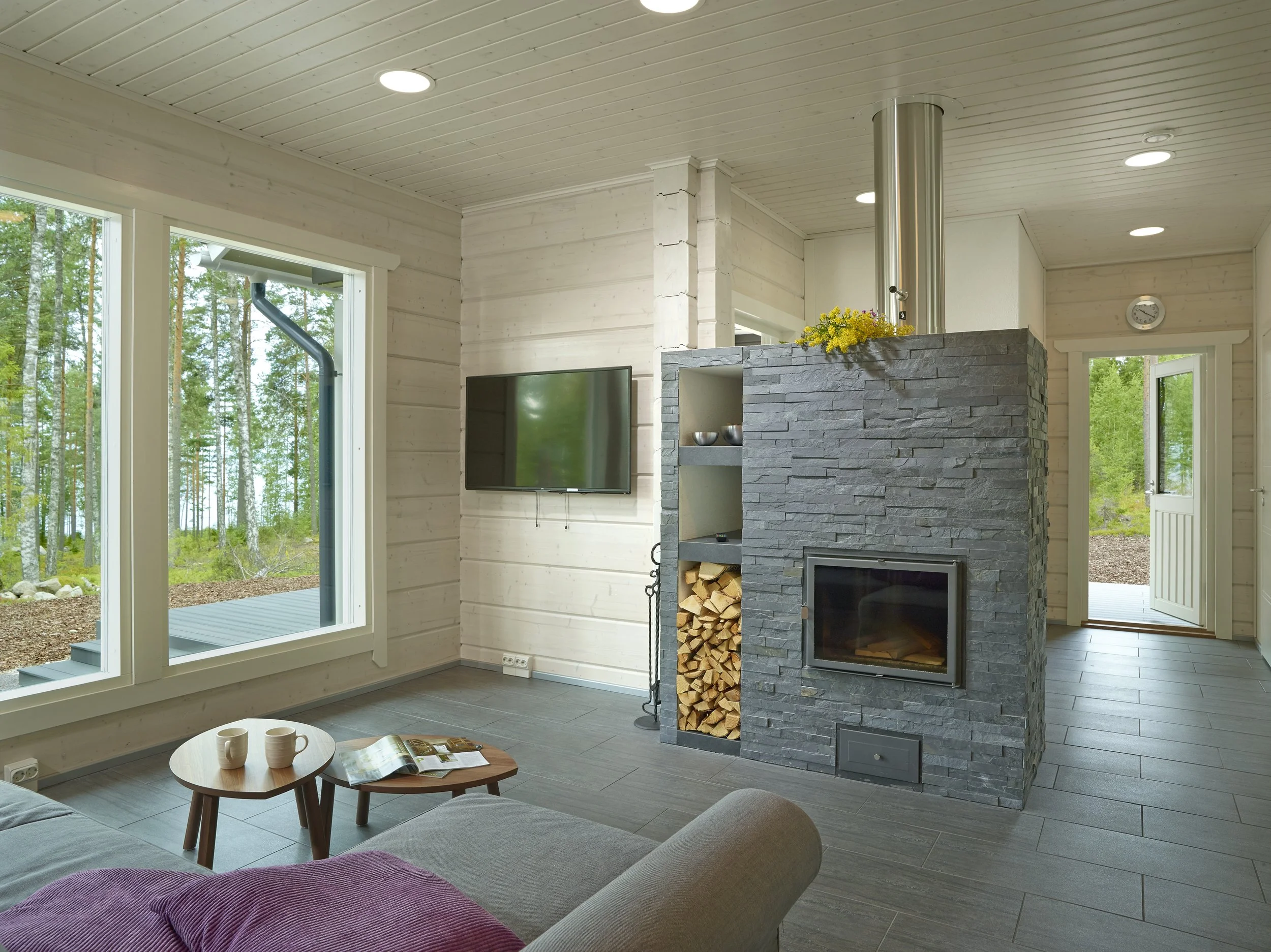 Living room with large window overlooking trees, modern fireplace with stacked firewood, mounted flat-screen TV, and seating area with mug and magazine.