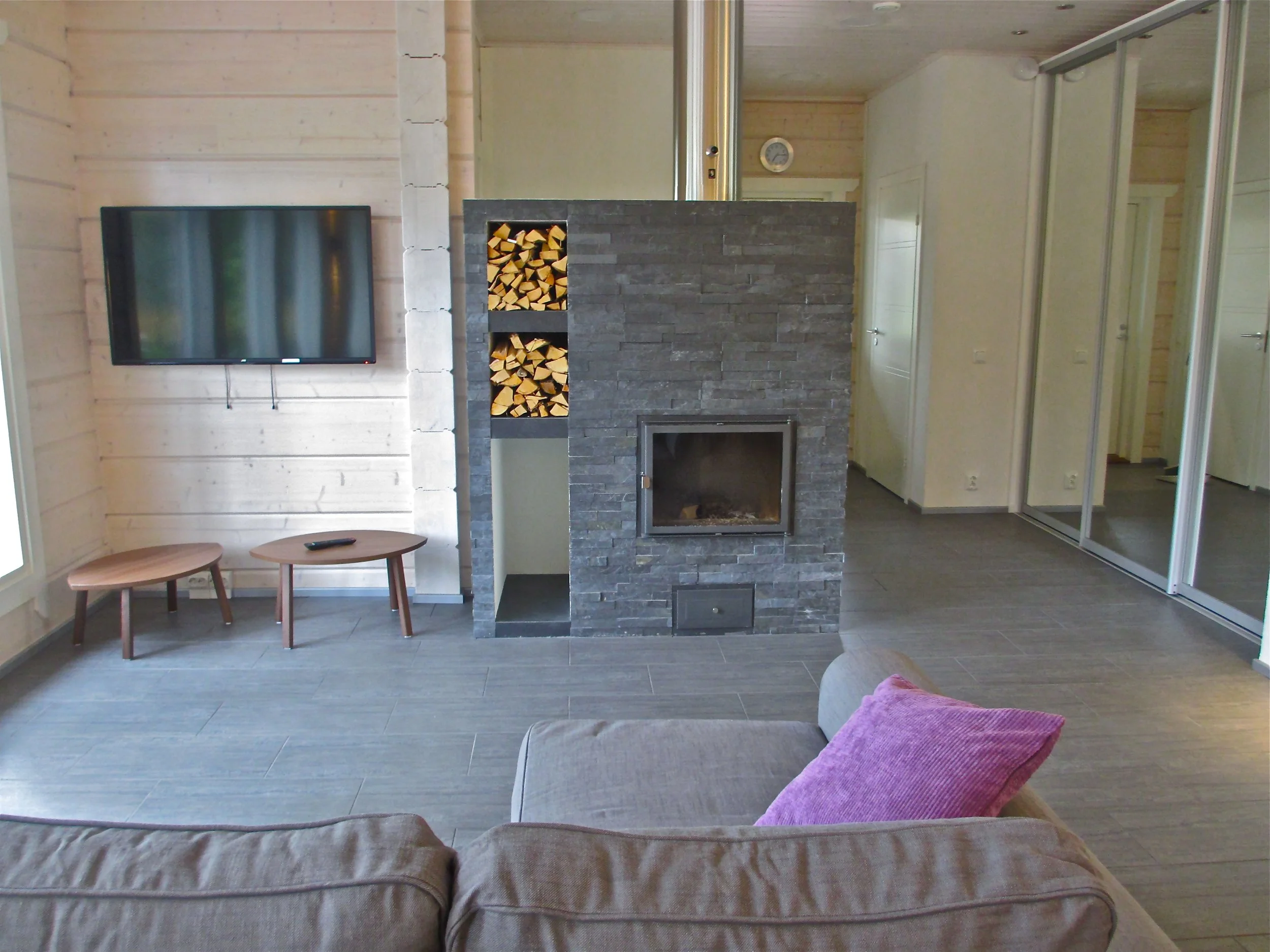 Living room with a wall-mounted TV, a fireplace with stacked wood, a beige sofa with a pink cushion, small wooden tables, and mirrored closet doors.