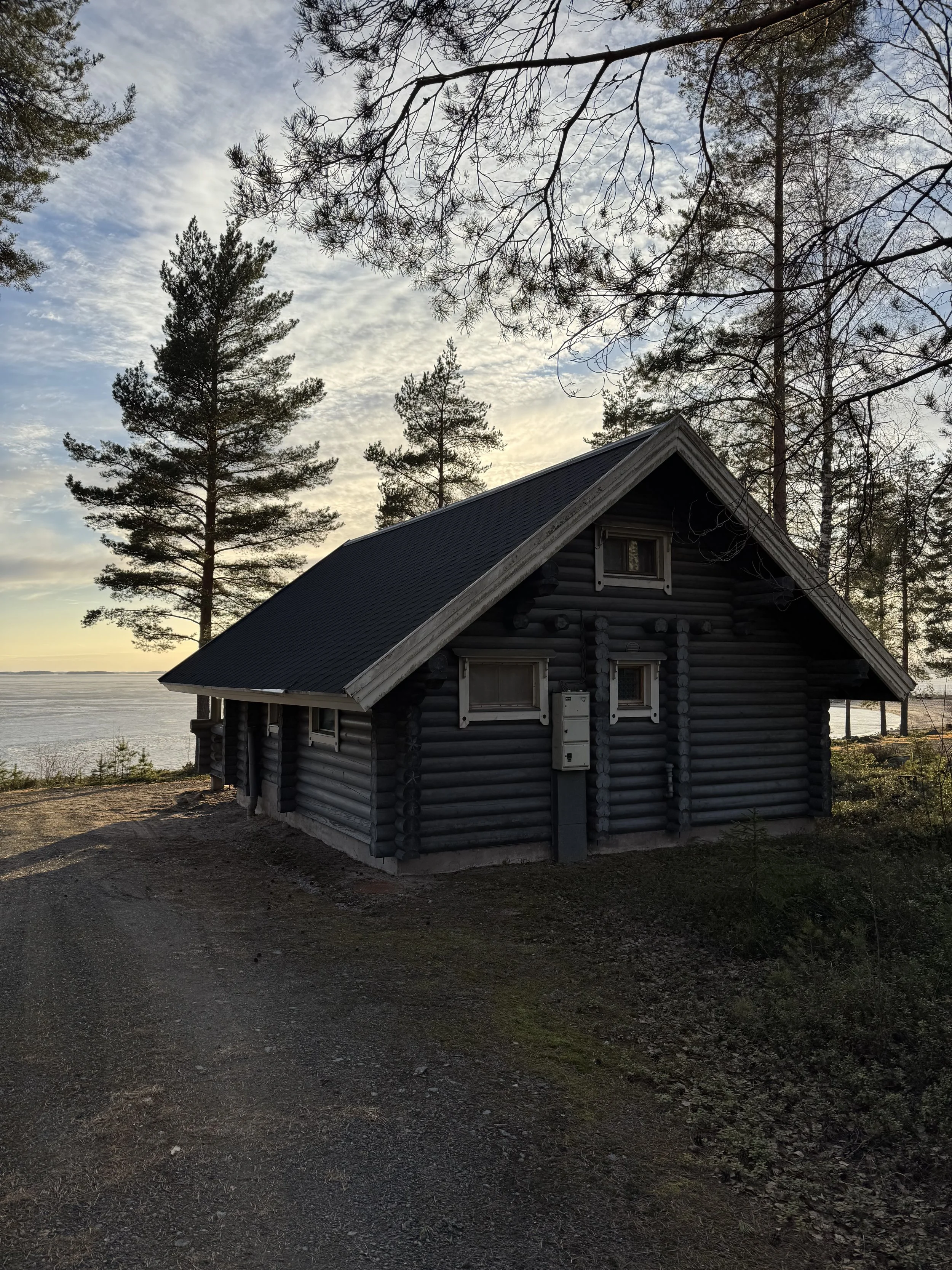 Pieni tumma hirsimökki, jossa on viisto katto, puiden ympäröimänä vesistön rannalla auringonlaskun aikaan.