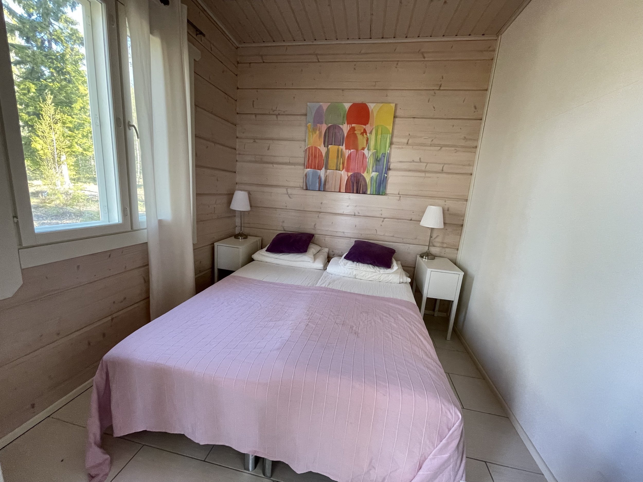 A bedroom with a bed covered in a pink quilt and two dark purple pillows. There are white nightstands with lamps on either side of the bed. A colorful abstract painting hangs above the bed. The wall behind the bed is wood-paneled, and there is a wind