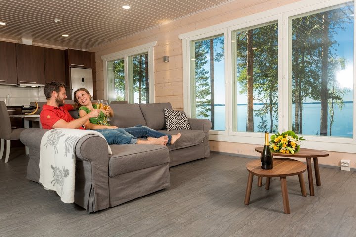 A couple sitting on a gray couch in a living room with large windows overlooking a lake. They are smiling and holding glasses, with a small round wooden table in front of them holding a bottle, a glass, and a floral arrangement.