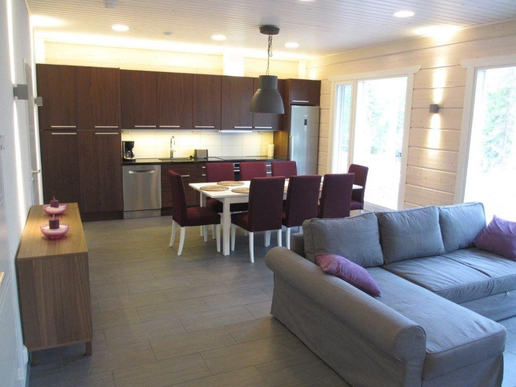 Open-plan living and dining area with a kitchen in the background, a gray sofa with purple pillows in the foreground, a wooden sideboard with pink decor, and large windows letting in natural light.
