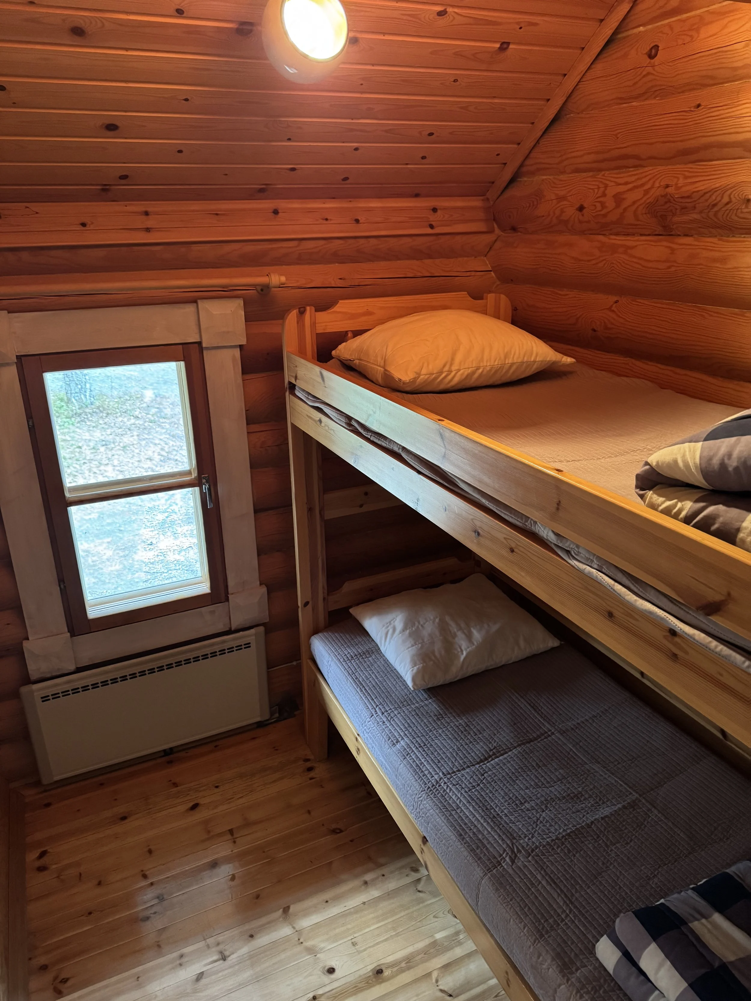 Viihtyisä puinen makuuhuone, jossa on kerrossänky ja pieni ikkuna; seinät, katto ja lattia ovat puuta, ja valo on päällä.