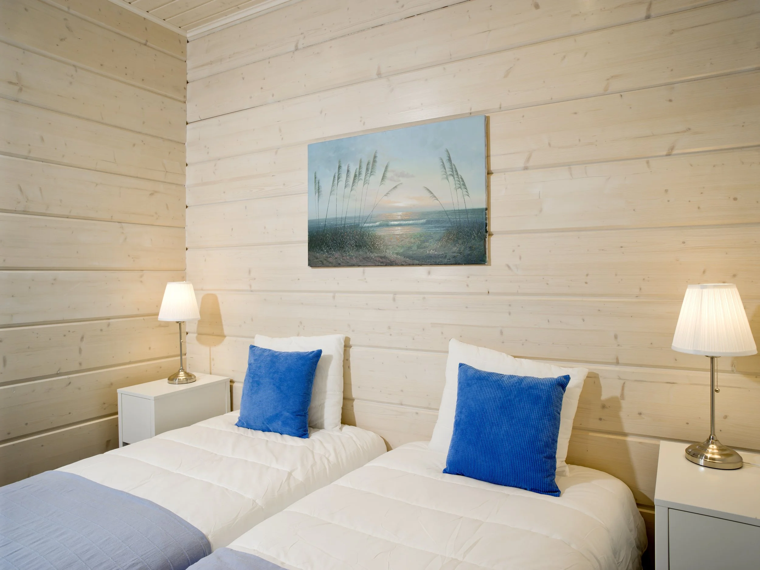 A bedroom with two twin beds, each with white bedding and a blue pillow, a nightstand with a lamp on either side, wooden walls, and a painting of a beach scene hanging above the beds.