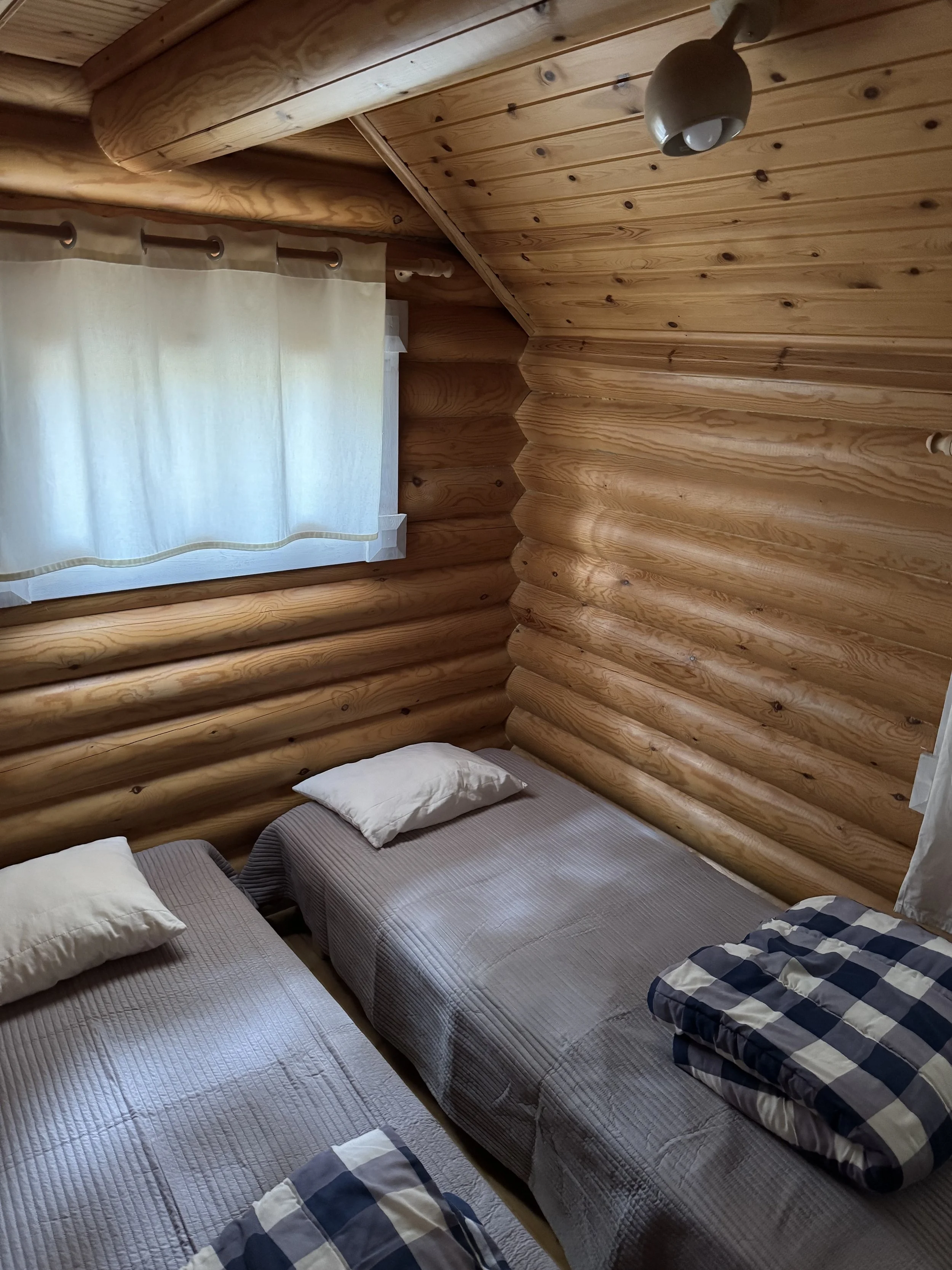 Kaksi sänkyä, joissa on tyynyt ja taitetut peitot, hirsimökin makuuhuoneessa, jossa on puiset seinät ja katto sekä ikkuna, jossa on valkoiset verhot.
