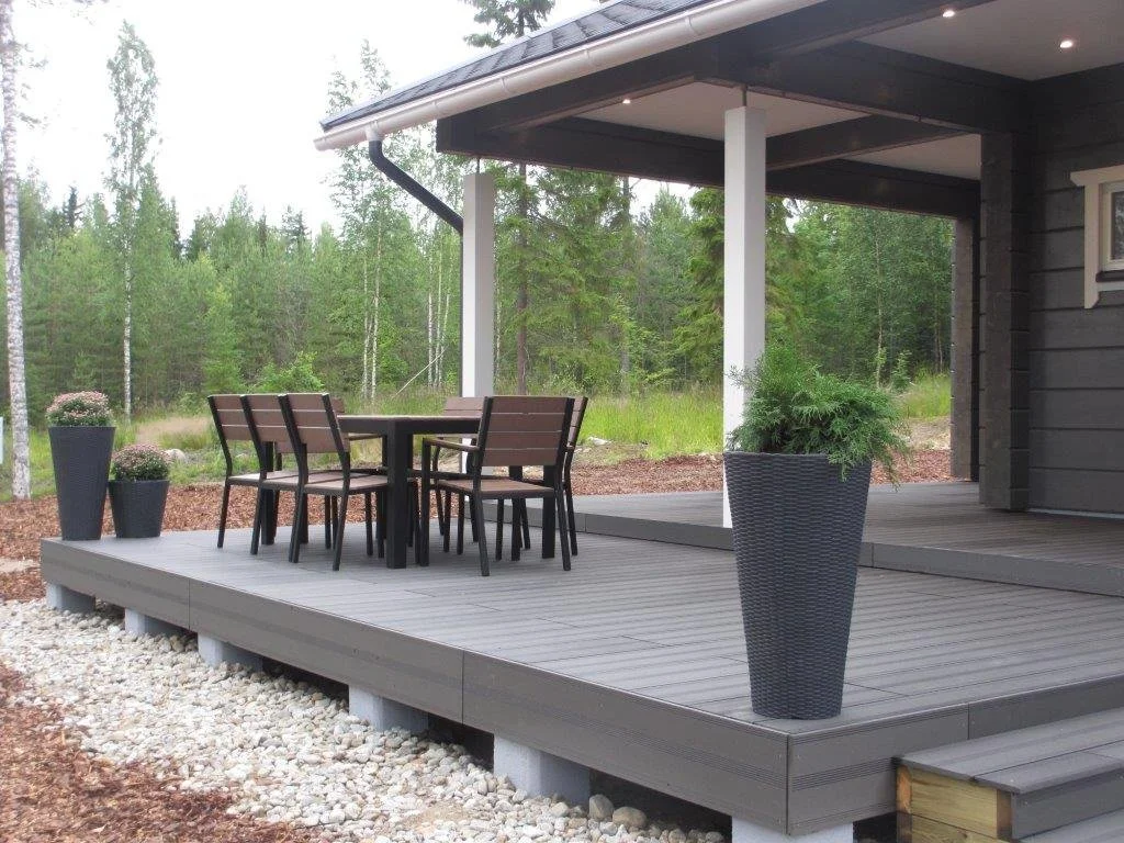 A backyard deck with a black dining table and four chairs, surrounded by potted plants in large black pots, with a wooded area in the background.
