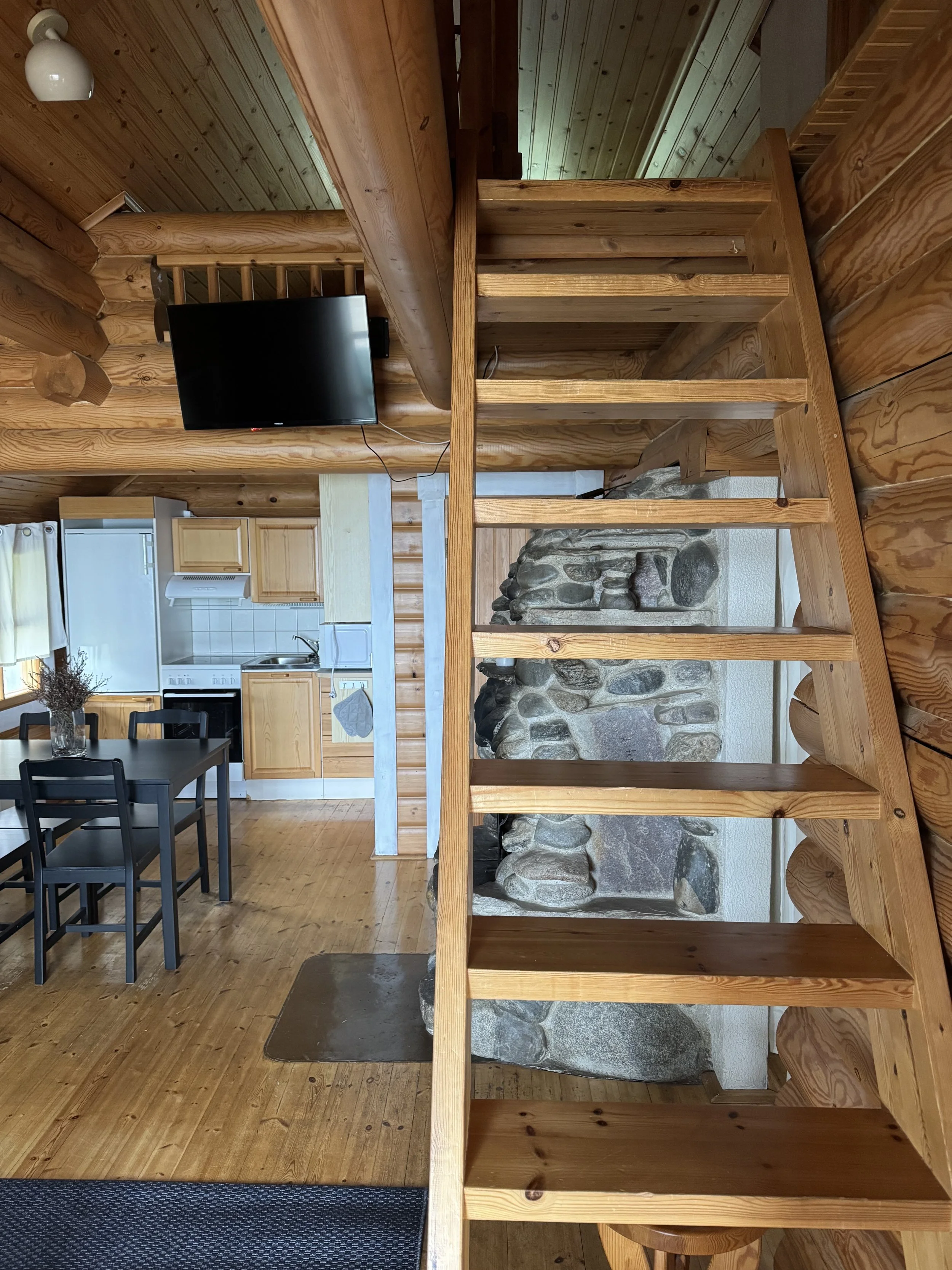 Hirsimökin sisätilat, joissa näkyy yläkertaan johtava portaikko, seinälle kiinnitetty taulutelevisio, ruokapöytä neljällä tuolilla, pieni keittiö vaaleilla puukaapistoilla ja jääkaappi sekä portaikon takana näkyvä kivitakka.