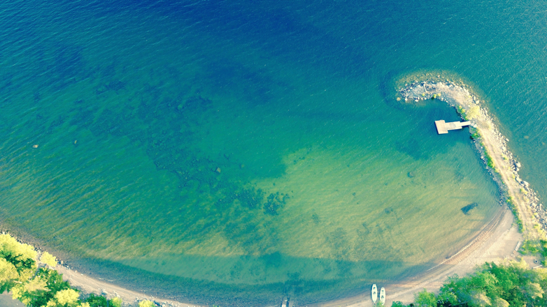 Ilmakuva Puruvesijärven pienestä rannasta, jossa vesi on kirkkaan sinistä ja vihreää, veteen ulottuva laituri sekä kaareva rantaviiva, jonka hiekalla on veneitä.