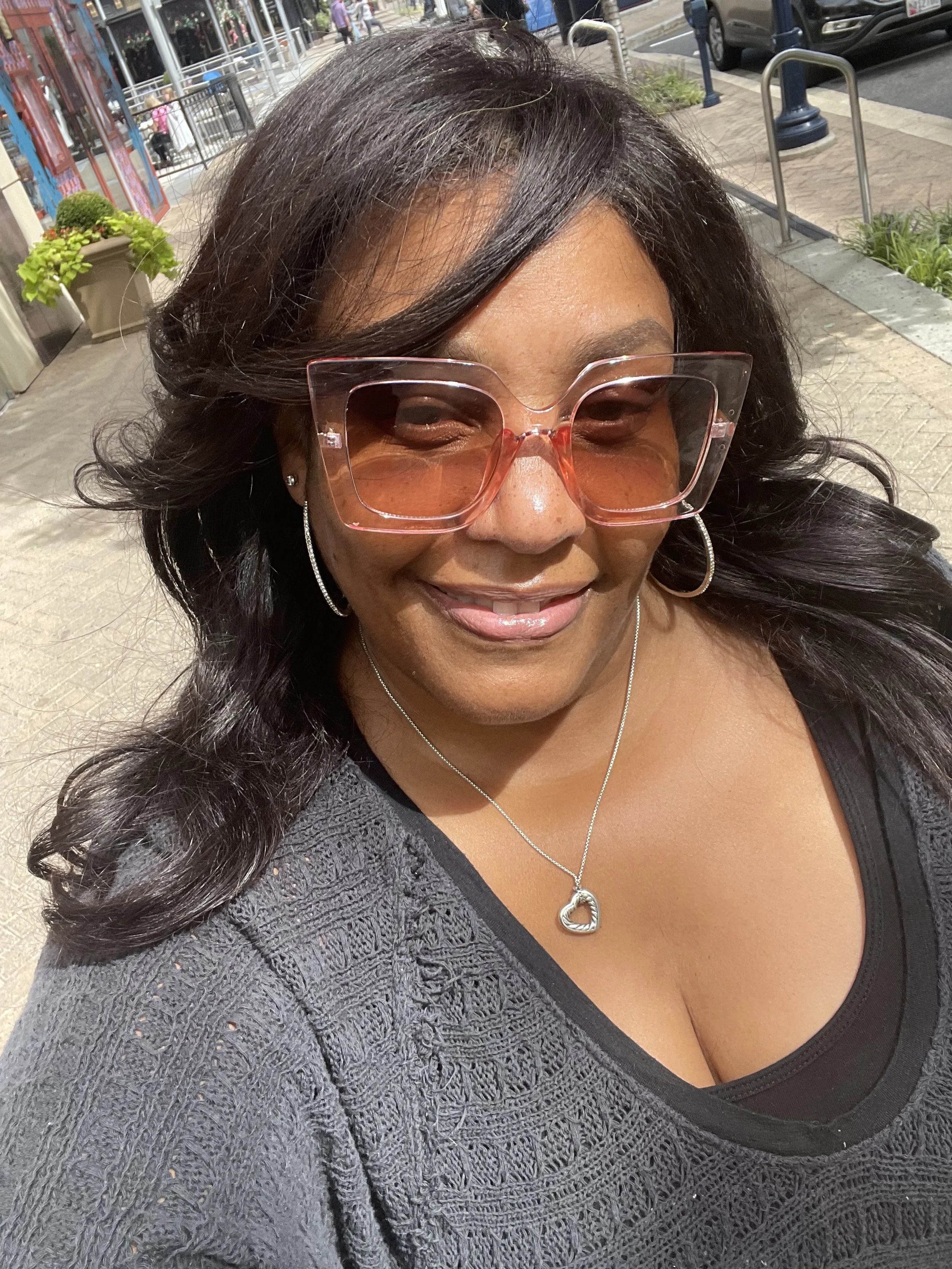 A woman with long, dark, wavy hair smiles while taking a selfie outdoors. She wears large, pink tinted sunglasses, silver hoop earrings, a silver heart-shaped necklace, and a dark, textured top with a black shirt underneath. The background shows a sidewalk, plants, and parked cars.