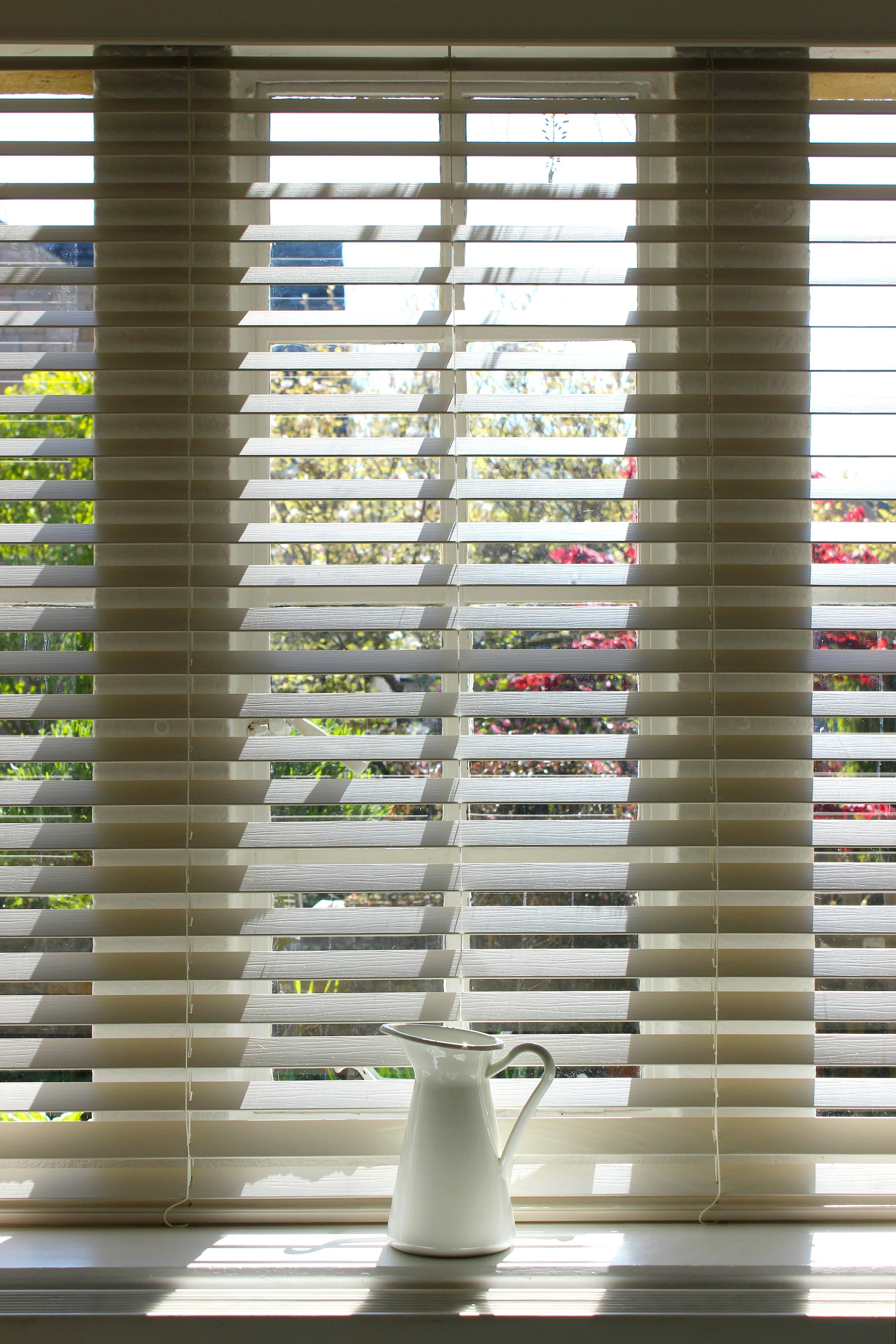 A white ceramic pitcher on a windowsill in front of closed white blinds, with a garden visible outside including green plants and blooming red flowers.