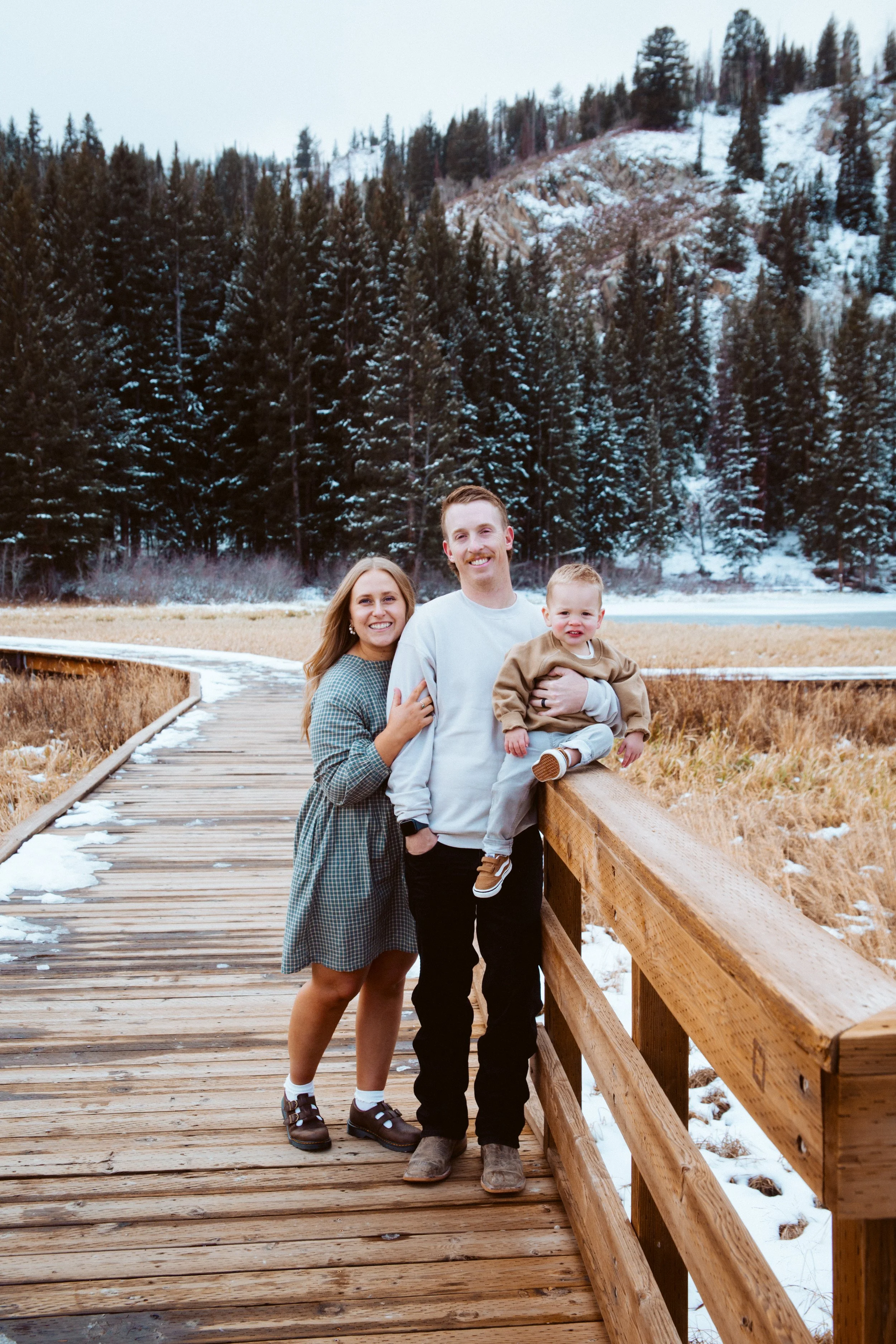 A family of three standing on a wooden walkway in a snowy, mountainous landscape with evergreen trees in the background.