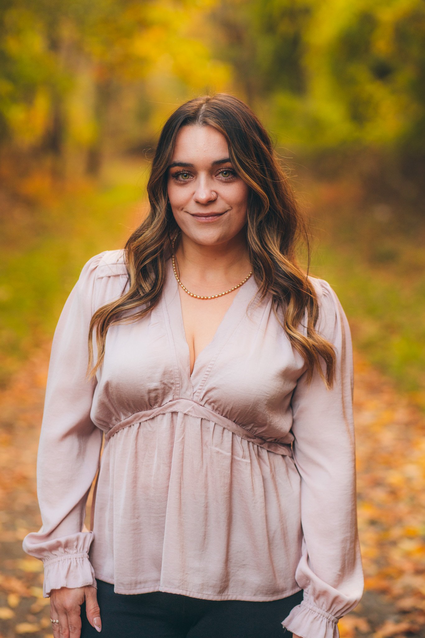 A woman with wavy brown hair and blue eyes standing outdoors on an autumn day with trees in the background. She is wearing a light pink blouse and a gold beaded necklace.