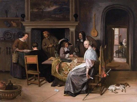 A group of six people, including women, men, and children, are gathered around a table in a room with a fireplace, paintings, and musical instruments. A woman in a white dress sits on a chair near the table. A dog is lying on the floor nearby.