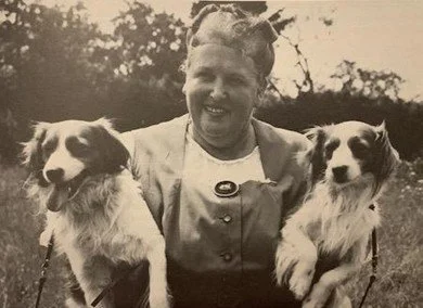 Mrs. M.C.S. Baroness van Hardenbroek van Ammerstol outdoors holding two dogs, one in each arm.