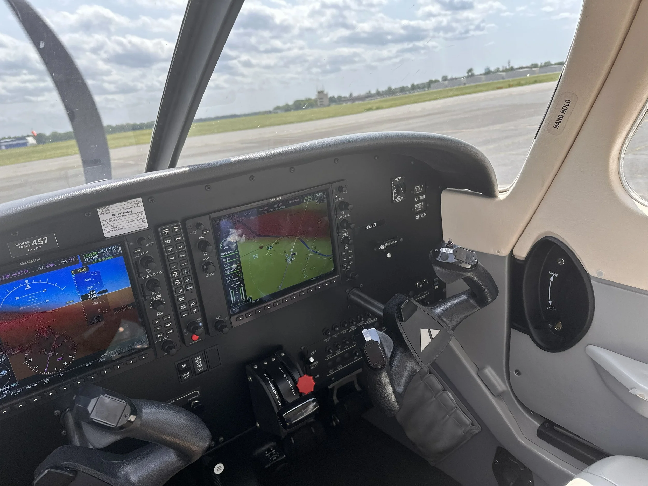 The cockpit of a small airplane showing various flight instruments and displays, with the view through the windshield of a runway, grassy area, and cloudy sky outside.
