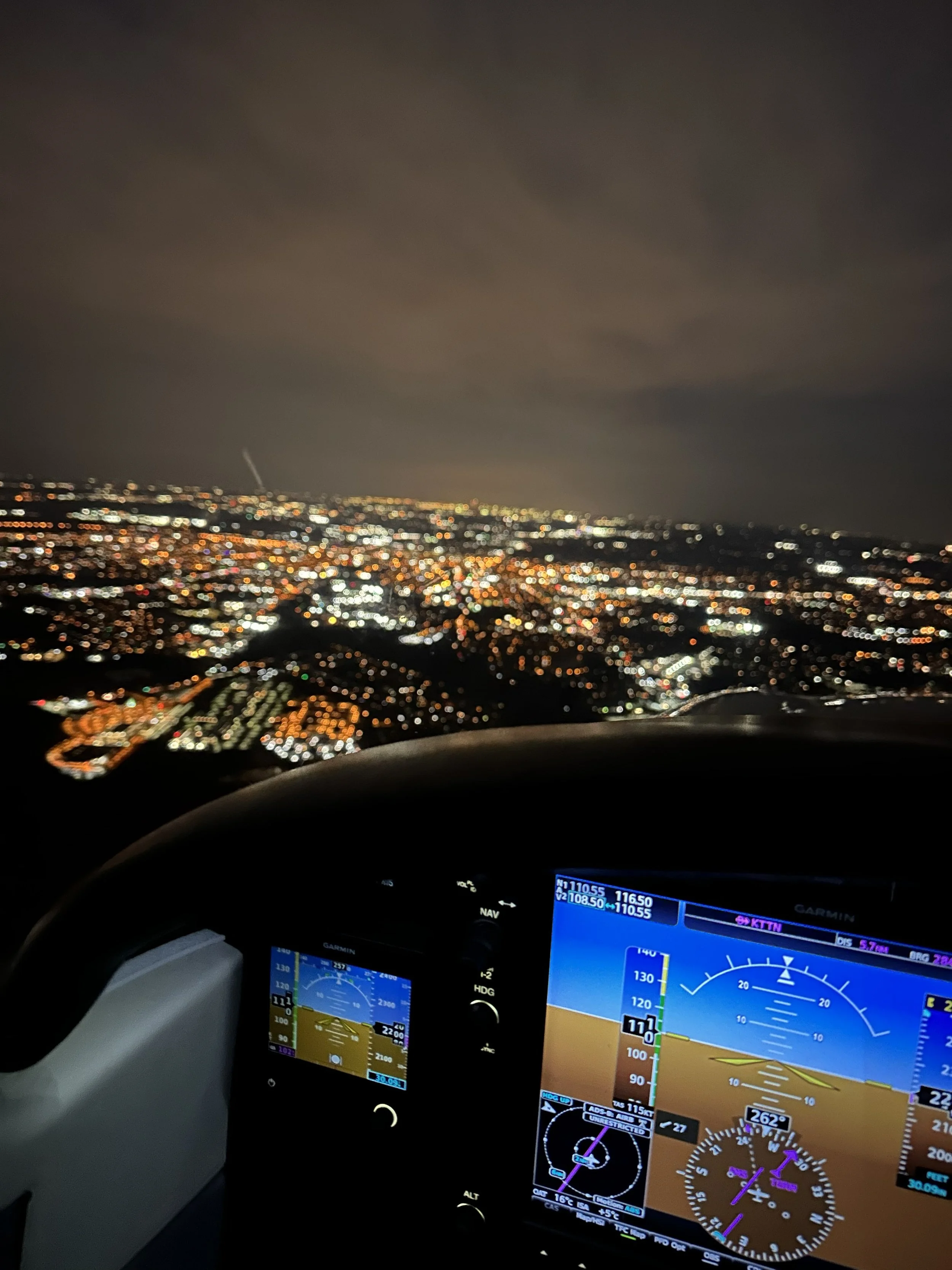 A nighttime view of a cityscape with illuminated buildings and streets, seen from inside an airplane cockpit. The cockpit displays aircraft instruments and navigation screens.