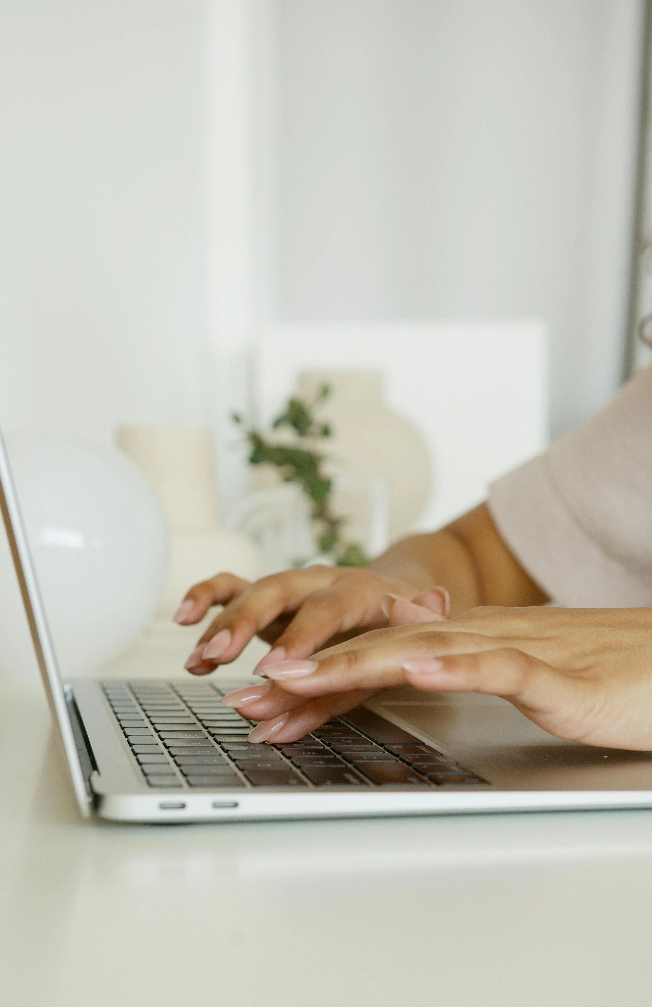 Close-up of hands typing on a silver laptop keyboard on a white desk; blurred background with a white wall, a mirror reflecting the hands, and a plant.