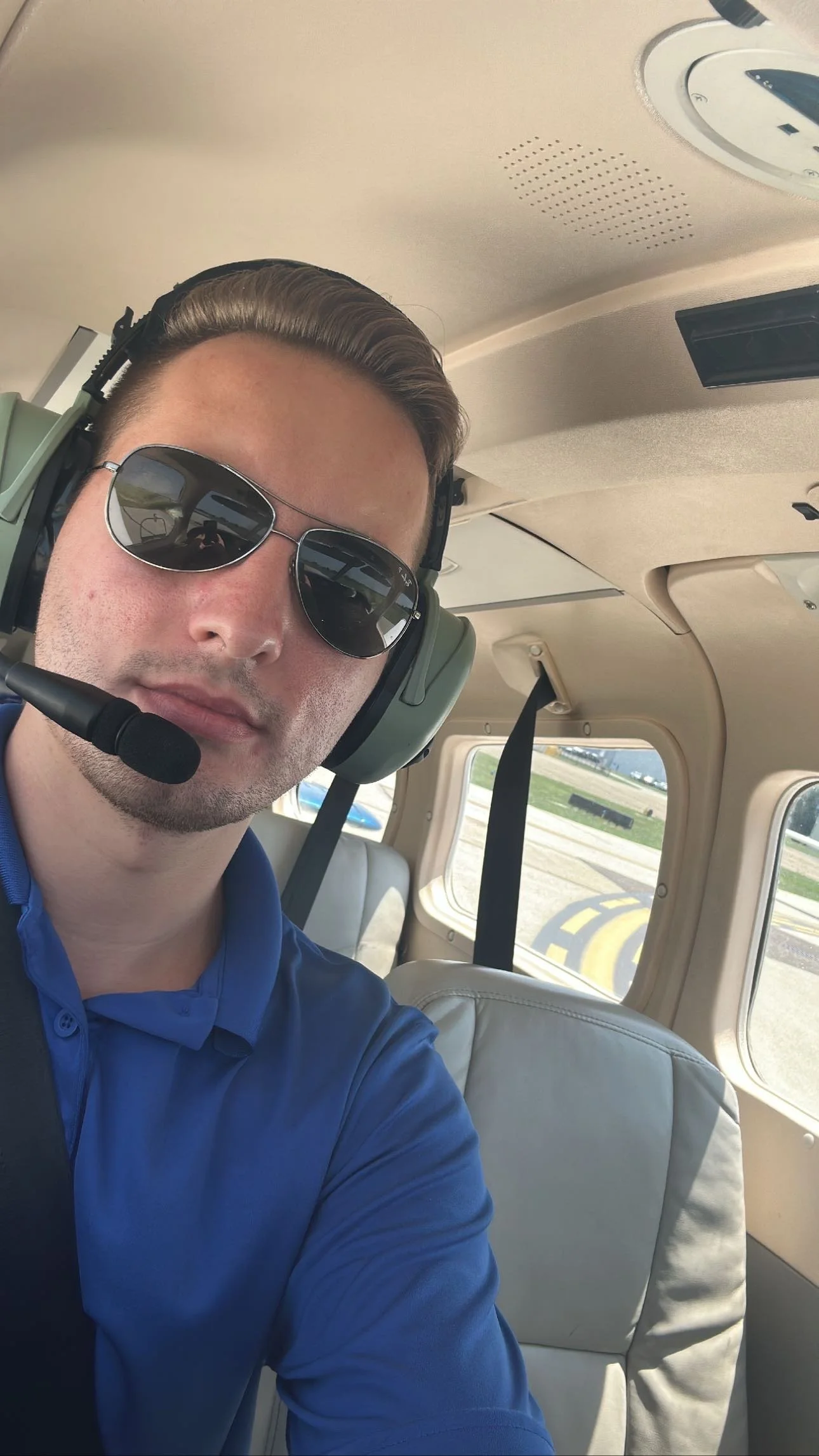 A man is sitting inside an aircraft, wearing sunglasses and a headset, and taking a selfie with part of his face visible. The aircraft's interior and runway can be seen through the window.