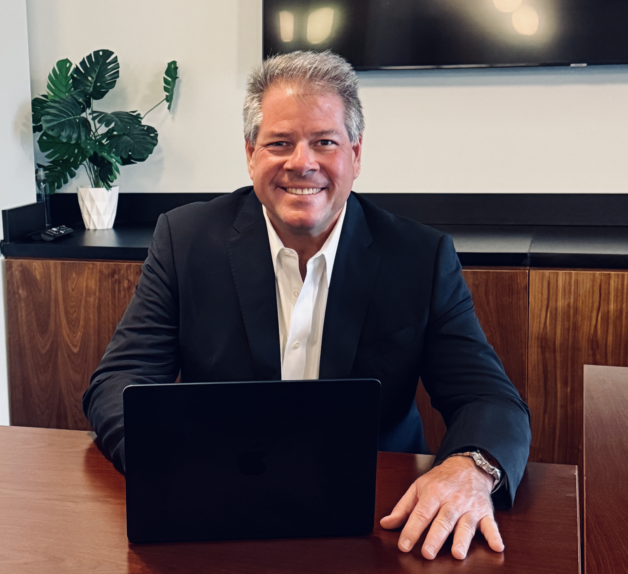 A lawyer in a black suit and white shirt sitting at a wooden table with a laptop in front of him, in a modern office setting with a large digital screen and a potted plant in the background.