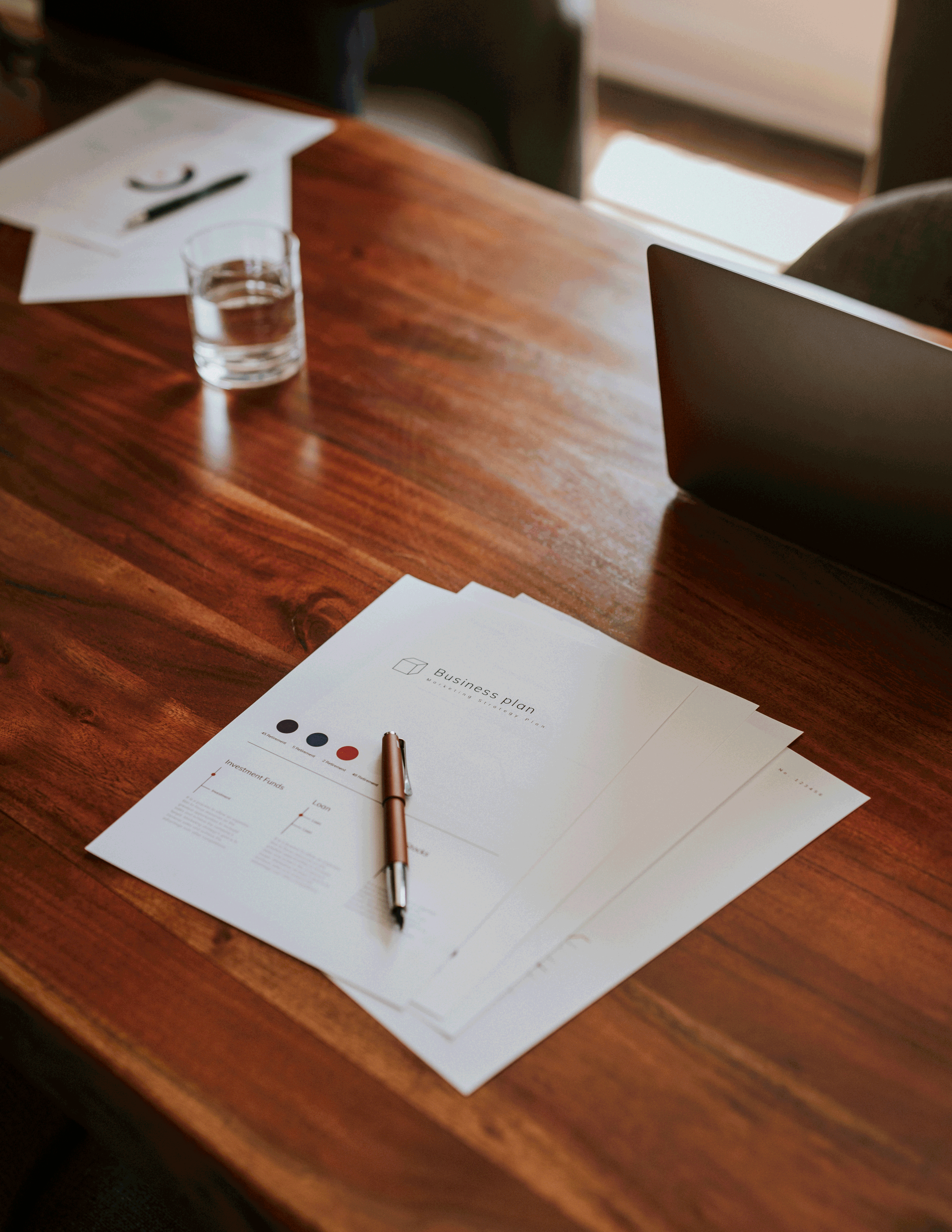A wooden table with papers, a pen, a glass of water, some papers, and a laptop.