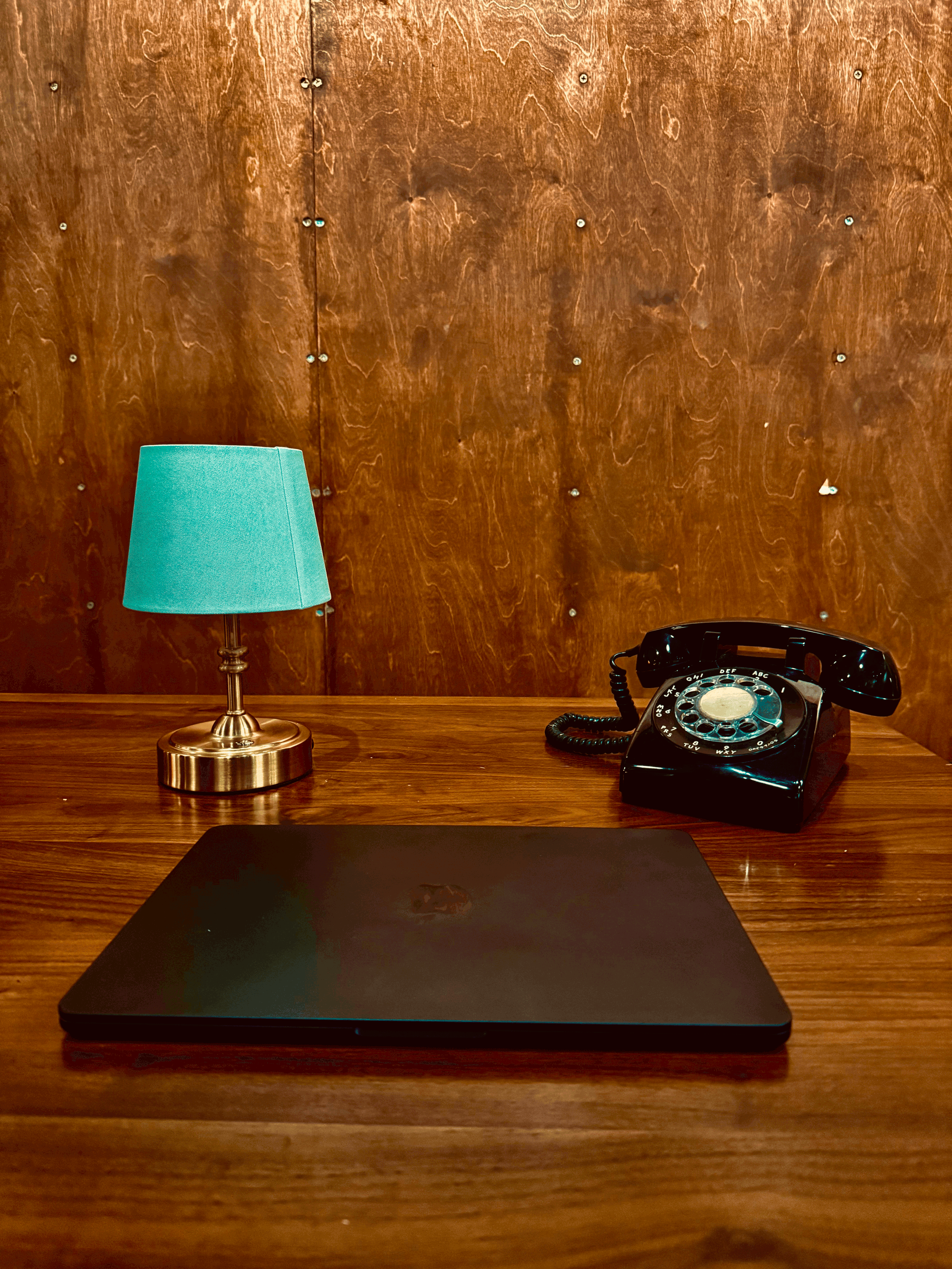 A wooden desk with a closed black laptop, a blue lampshade, and a black rotary dial telephone in front of a wooden panel wall.