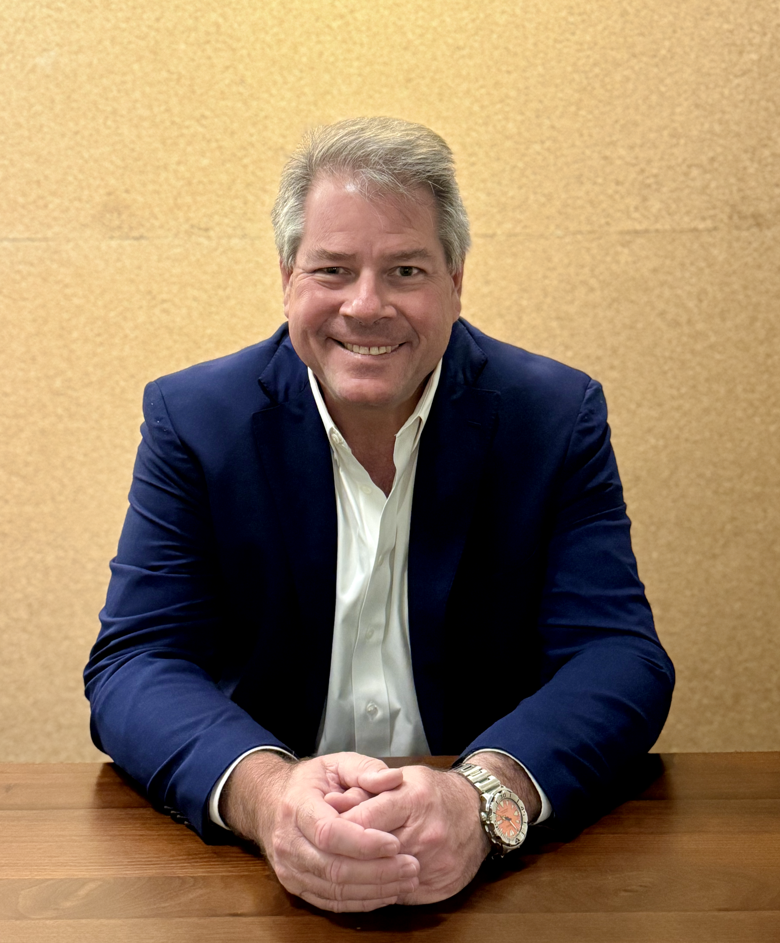 A man in a blue suit and white shirt sitting at a wooden table, smiling at the camera against a beige wall.