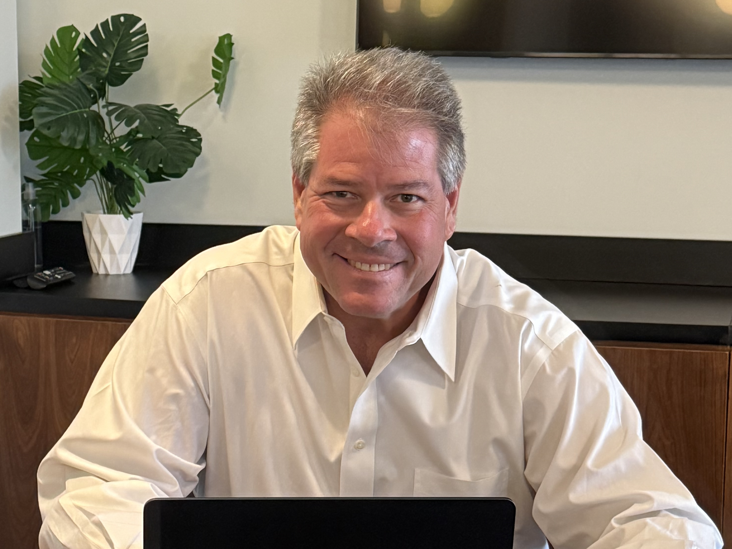 A smiling man with gray hair in a white shirt sitting at a table with a laptop in front of him. Behind him is a green monstera plant in a white pot and a wooden cabinet.