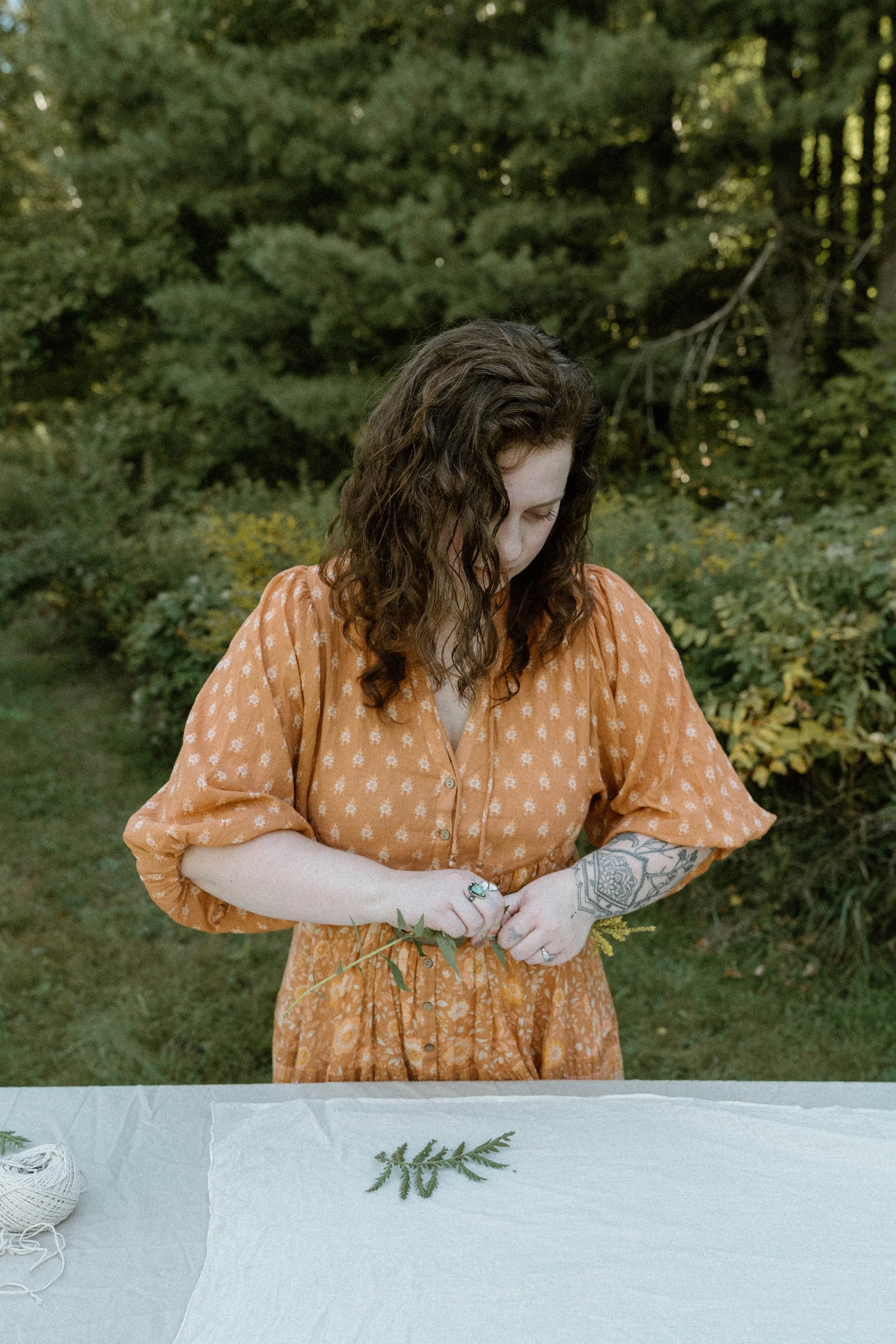 A woman with curly hair wearing an orange dress with white floral patterns is standing outdoors in a wooded area, holding green foliage.