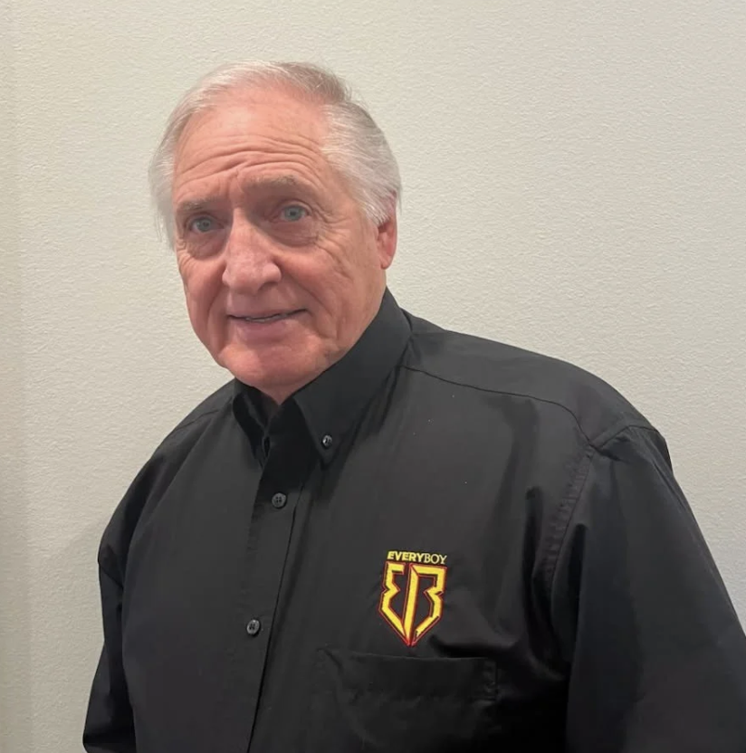 An elderly man with gray hair wearing a black collared shirt with an embroidered logo that says 'Everybody' and a shield emblem.